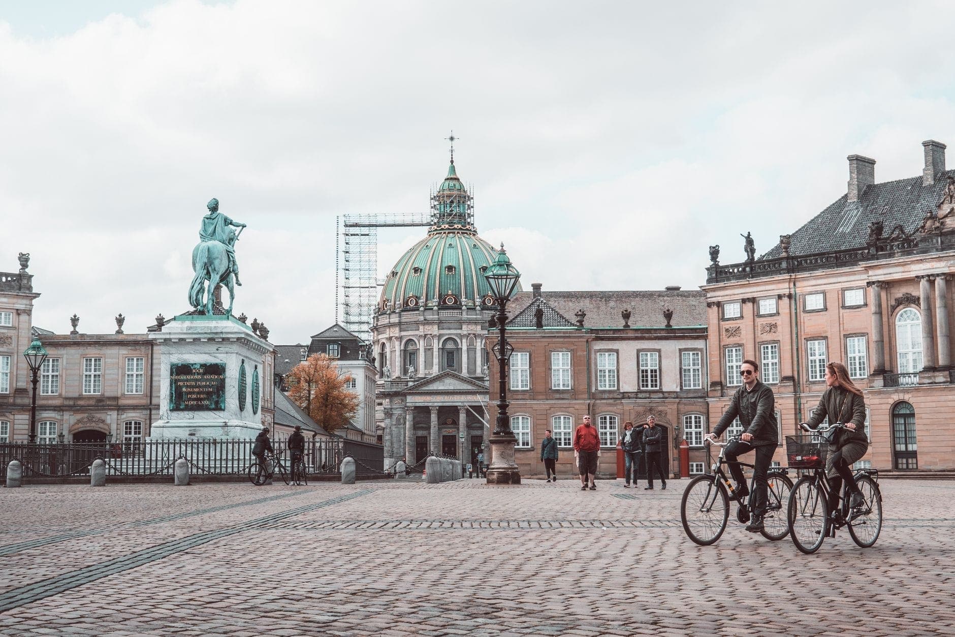 Zwei Menschen radeln durch einen offenen Stadtplatz in Kopenhagen mit der Marmorkirche und historischen Gebäuden im Hintergrund an einem bewölkten Tag.