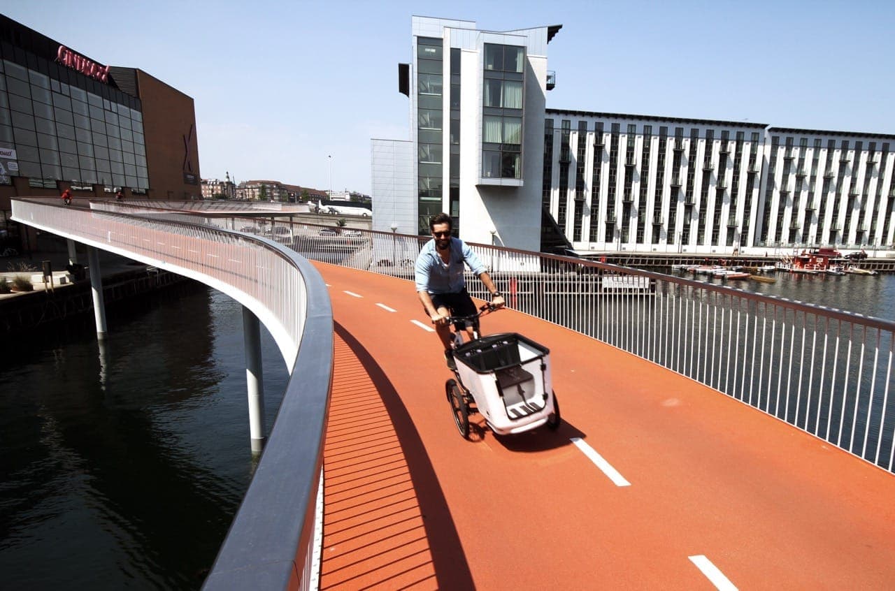 El puente ciclista Cykelslangen en Copenhague se curva sobre el puerto, con un ciclista recorriendo su sendero naranja brillante y edificios modernos al fondo.