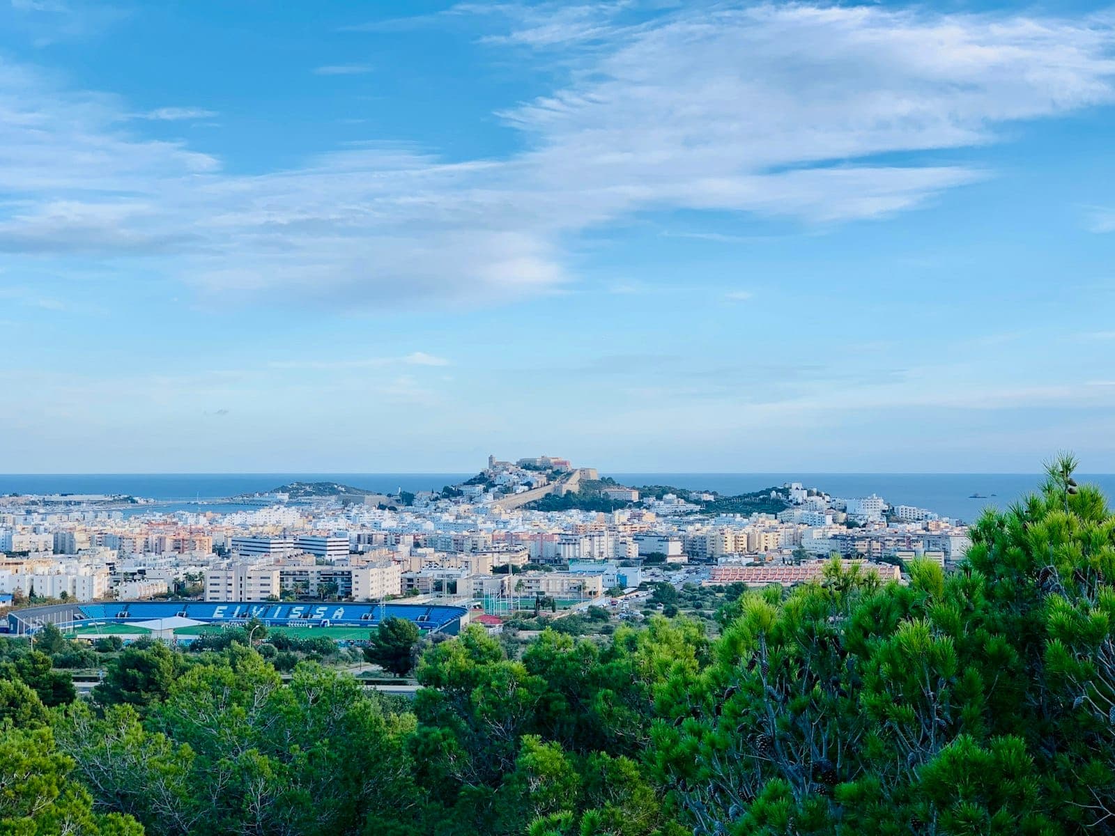 Panoramic view over Ibiza Town with Dalt Vila’s fortified old city rising above the modern town, surrounded by greenery and blue sky.