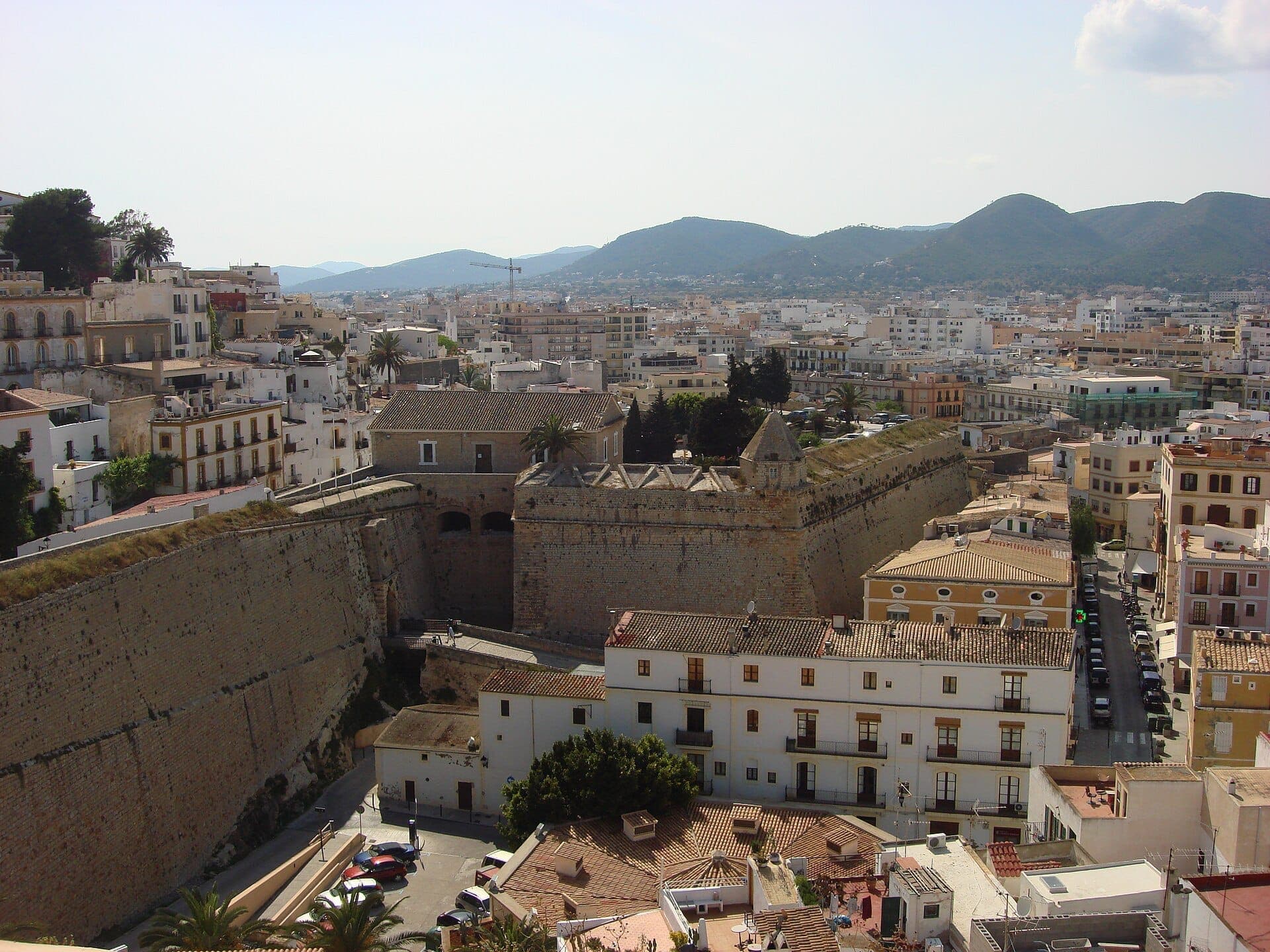 View of the massive stone Dalt Vila fortress walls surrounded by historic buildings in Ibiza Town, with mountains in the background.