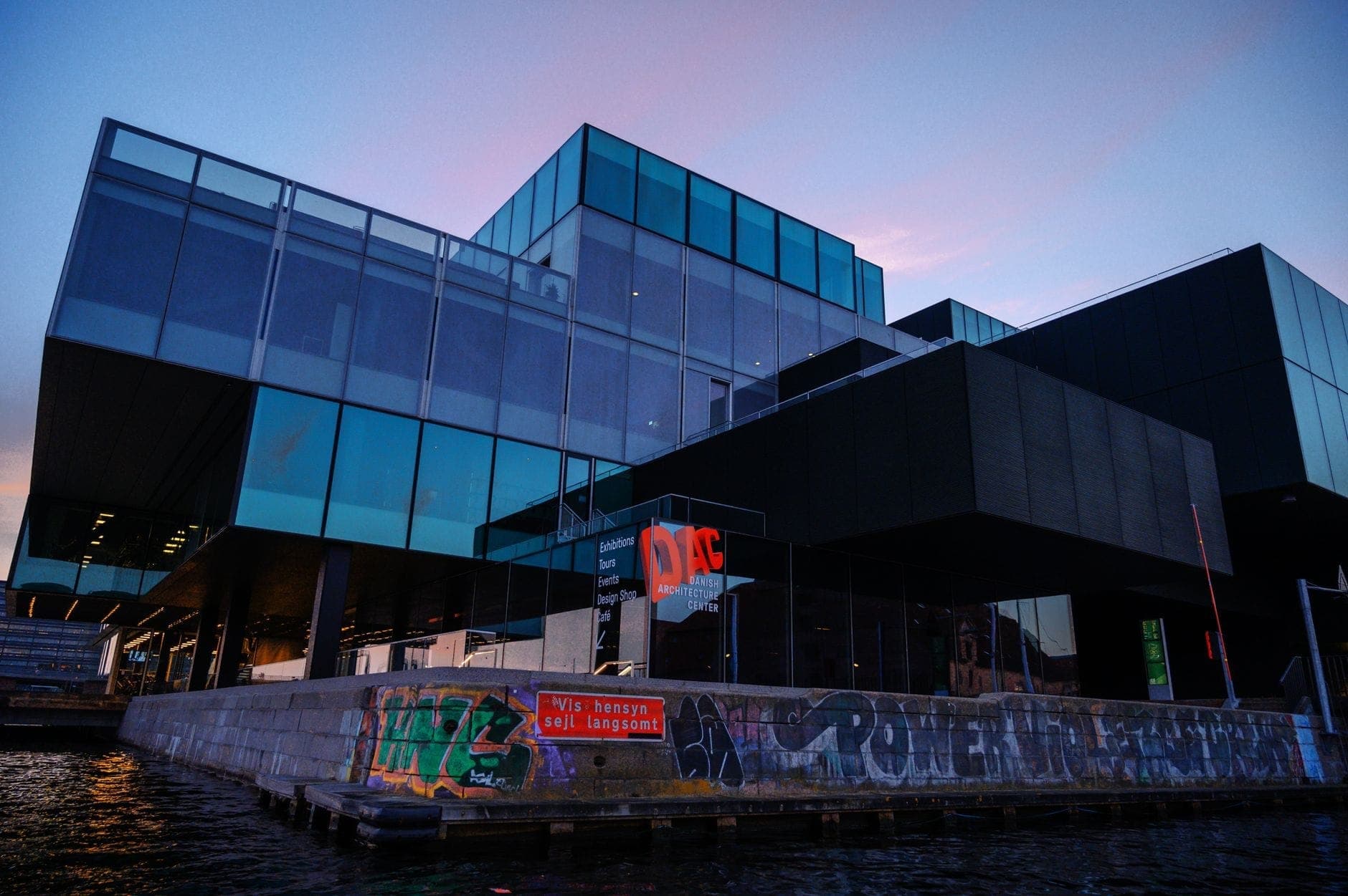 Moderne Glasarchitektur des BLOX-Gebäudes des Dänischen Architekturzentrums bei Sonnenuntergang, vom Hafenufer aus gesehen mit Spiegelungen und urbanem Graffiti.