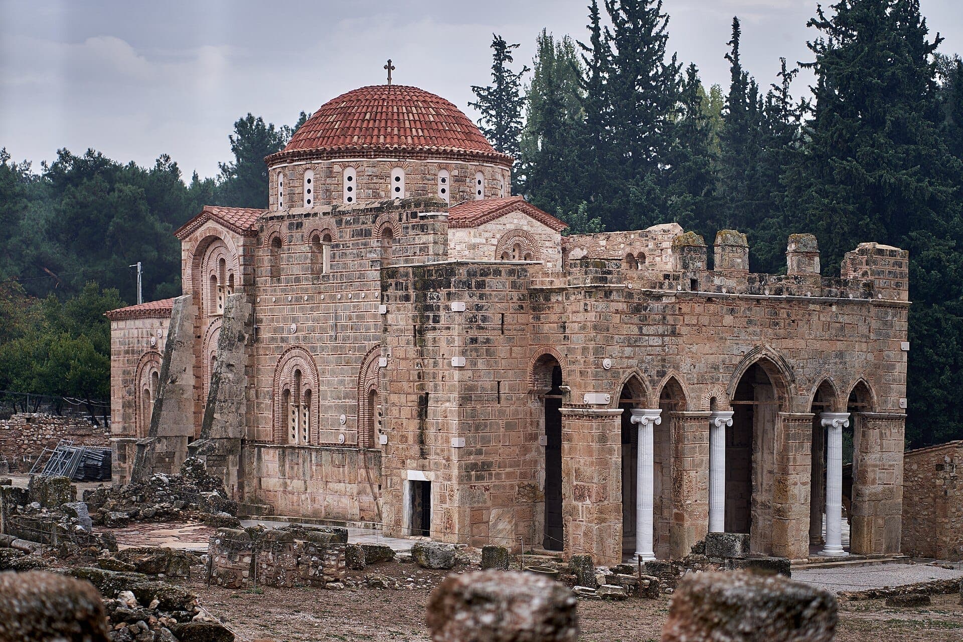 Daphni Monastery with its distinctive Byzantine dome, stone arches, and columns stands surrounded by greenery and ancient ruins under a cloudy sky.