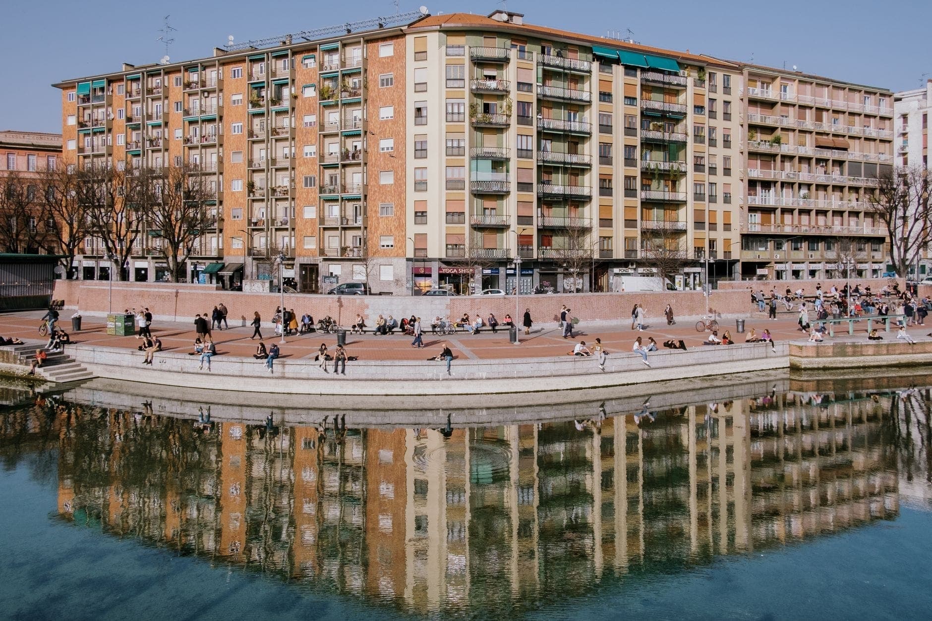People relax on the steps of Darsena di Milano, with colorful apartment buildings and their reflections visible in the calm waterfront basin.
