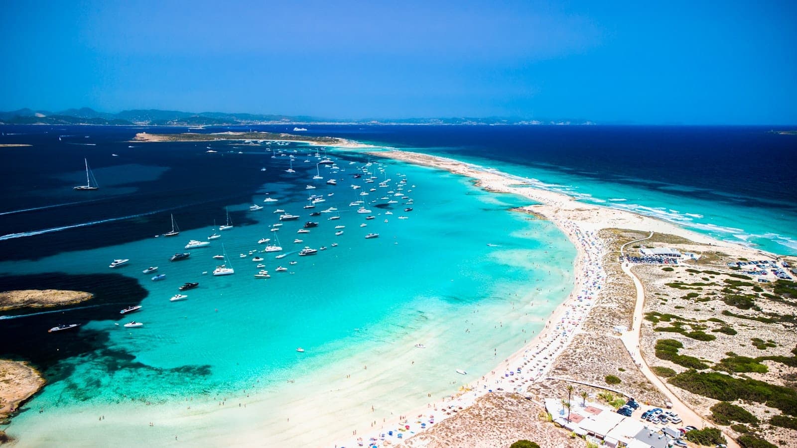 Aerial view of Formentera's turquoise beaches, sandbars, and many boats anchored in crystal-clear water under a bright blue sky.