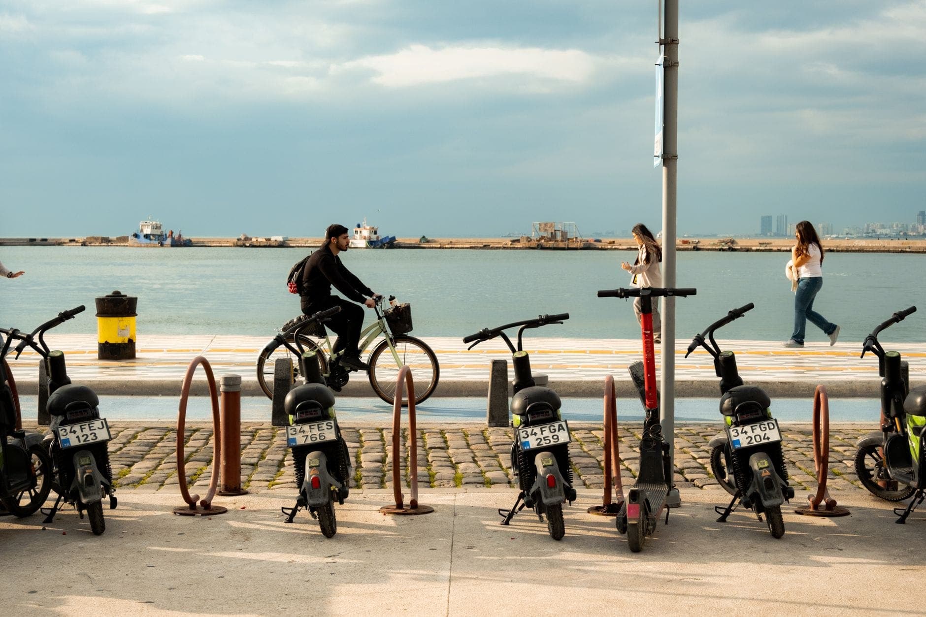 Port area with scooters and bikes parked, people walking and cycling along waterfront under cloudy sky in a Mediterranean setting.