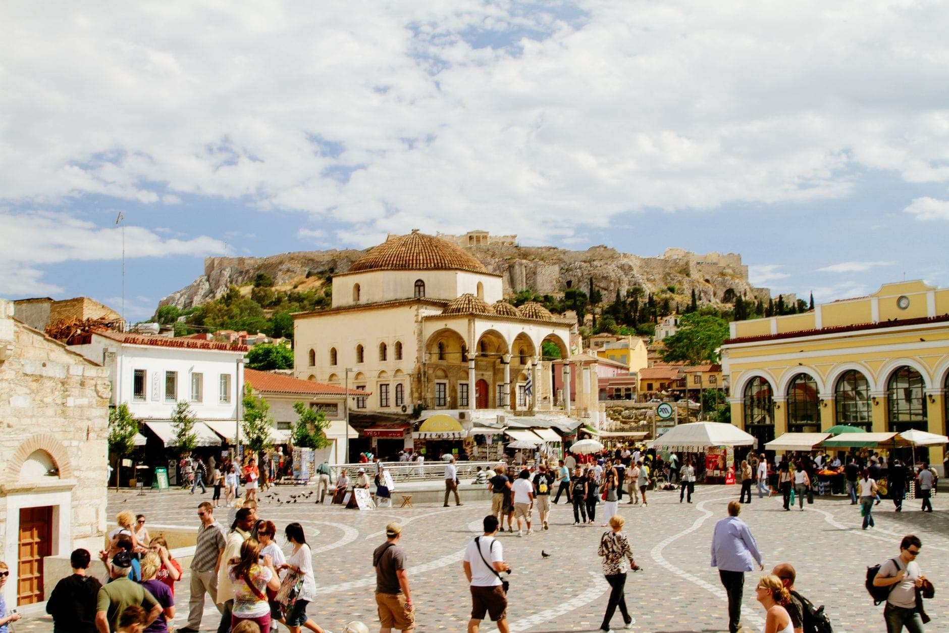 Belebter Platz in Athen mit Menschenmassen, einer historischen Moschee und der Akropolis im Hintergrund unter einem teilweise bewölkten Himmel.