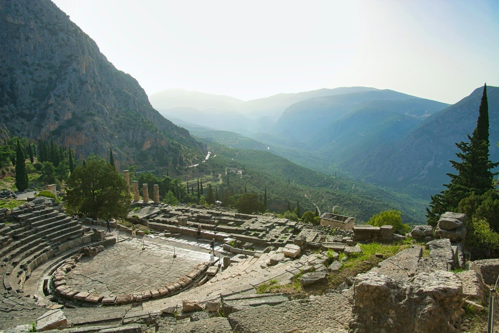 Panoramablick auf ein antikes griechisches Theater an einem Hang, umgeben von Bergen und Grün unter einem diesigen Himmel.