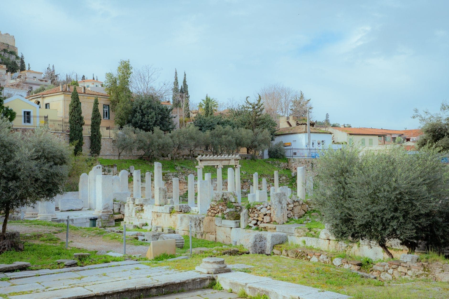 Steinruinen und Marmorsäulen einer antiken archäologischen Stätte in Athen, mit Grünflächen und Häusern im Hintergrund.