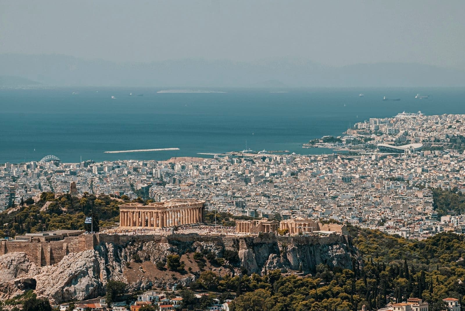 A wide aerial view of Athens showing the Acropolis and Parthenon with the city extending to the sparkling blue sea under clear skies.
