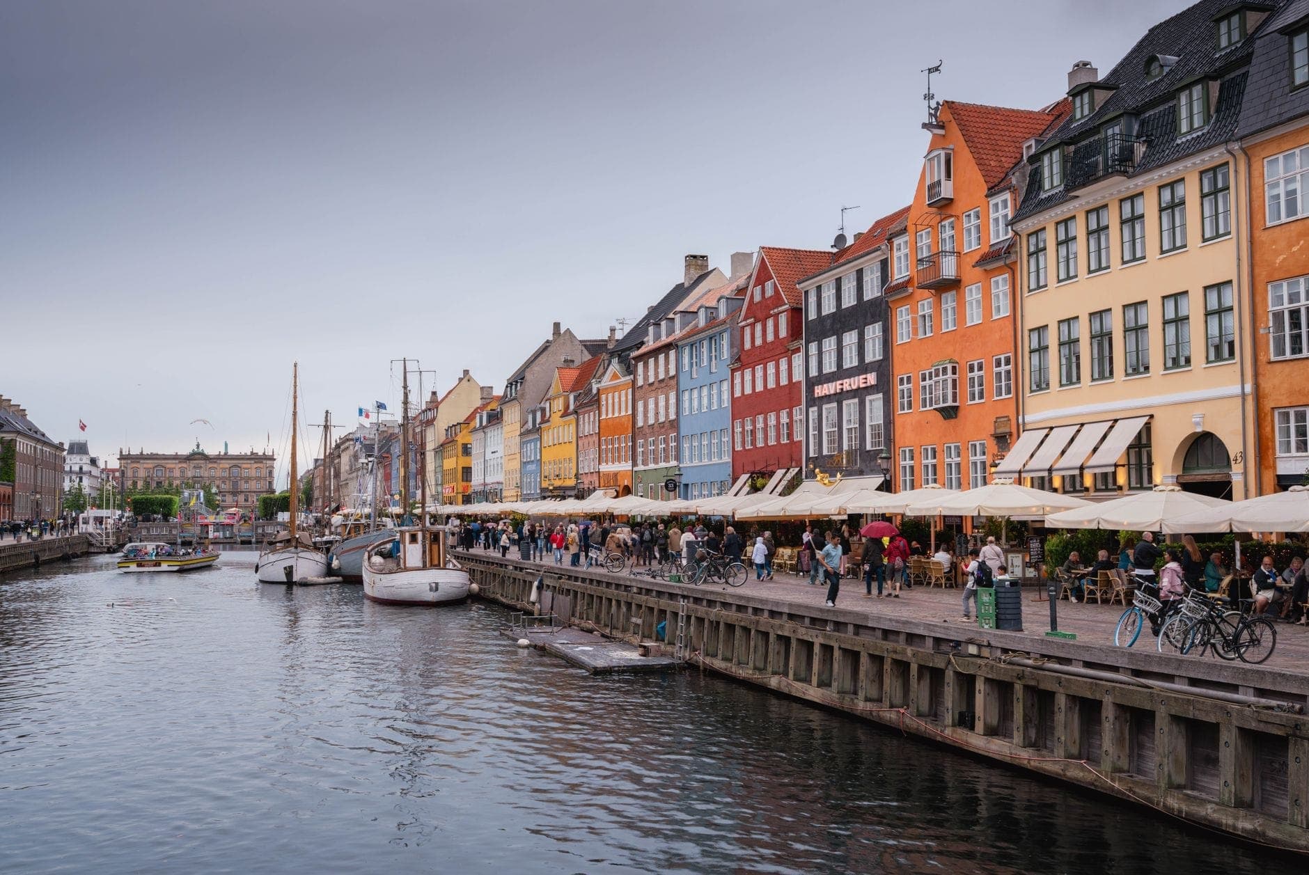 Vivace scena di canale a Copenaghen con edifici storici colorati, barche ormeggiate e persone che passeggiano sul lungofiume sotto un cielo nuvoloso.
