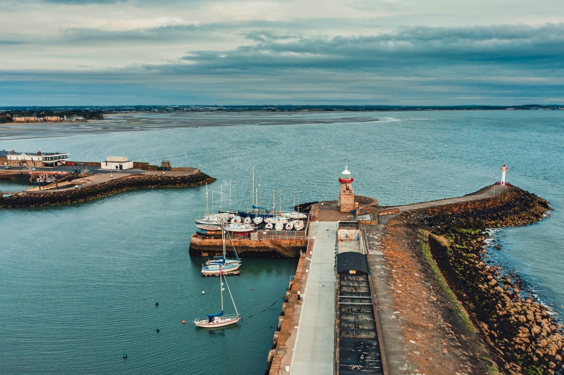 Vue aérienne d'un port avec de petits bateaux, une jetée en pierre et deux phares le long du littoral de Dublin sous un ciel nuageux.