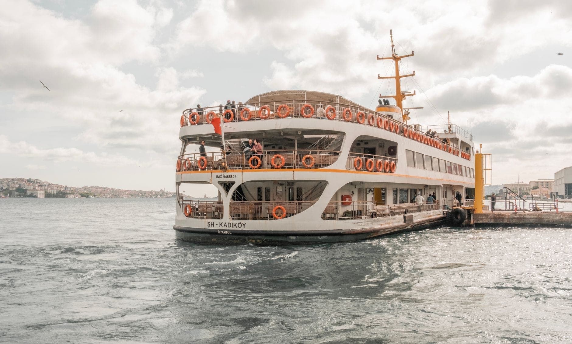 Duży prom w Stambule z napisem 'Kadıköy' zacumowany przy nabrzeżu, z widocznymi pasażerami na pokładzie i linią brzegową miasta w tle.