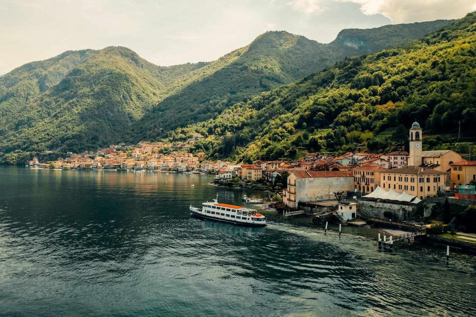 Traghetto in arrivo in un pittoresco paese sul lago con edifici dai tetti arancioni, immerso tra montagne verdi e le acque calme di un lago italiano.