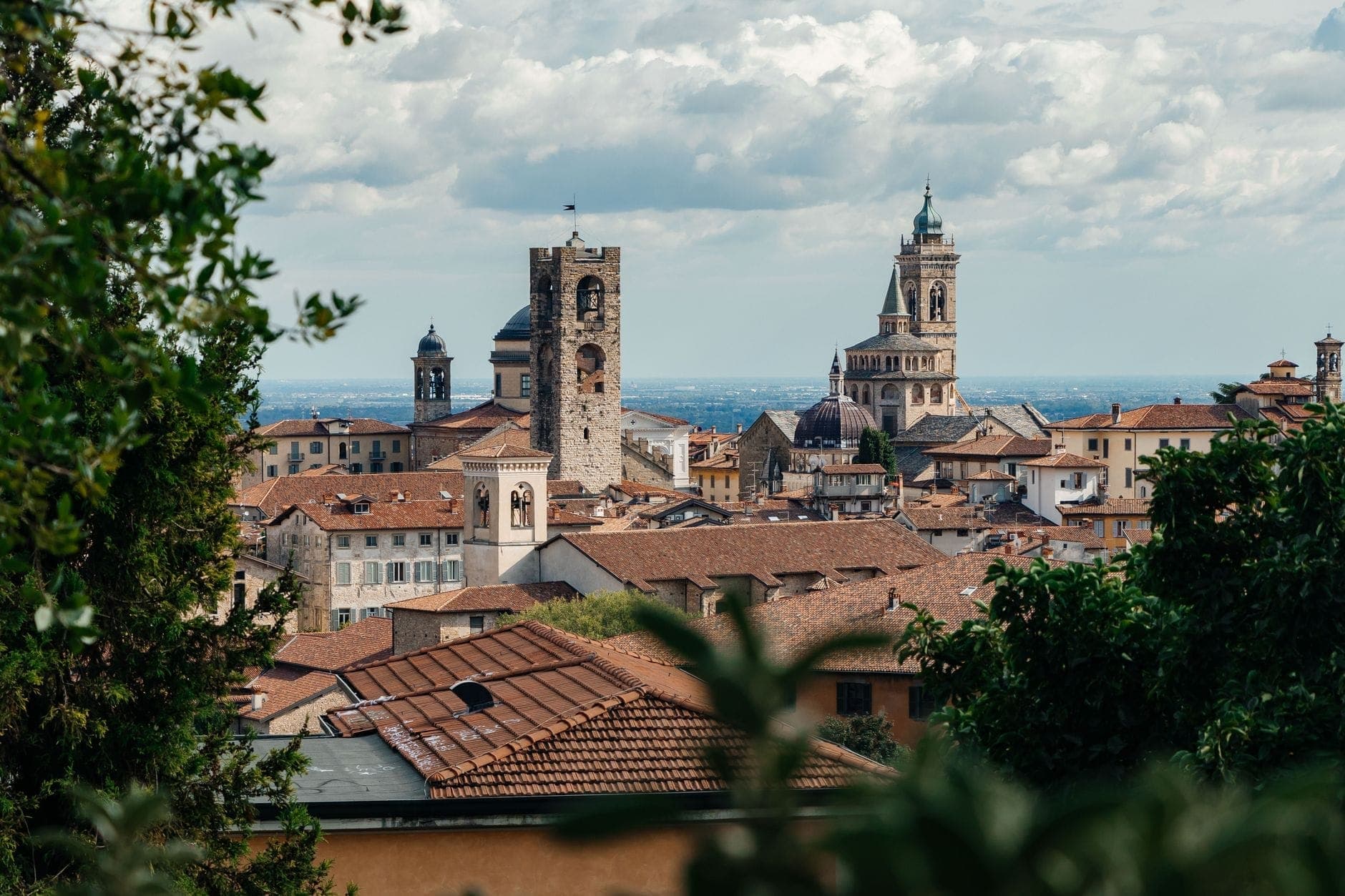 Vista panoramica dello skyline medievale di Bergamo con torri storiche e guglie di chiese che si innalzano sopra i tetti in terracotta, incorniciati dagli alberi.