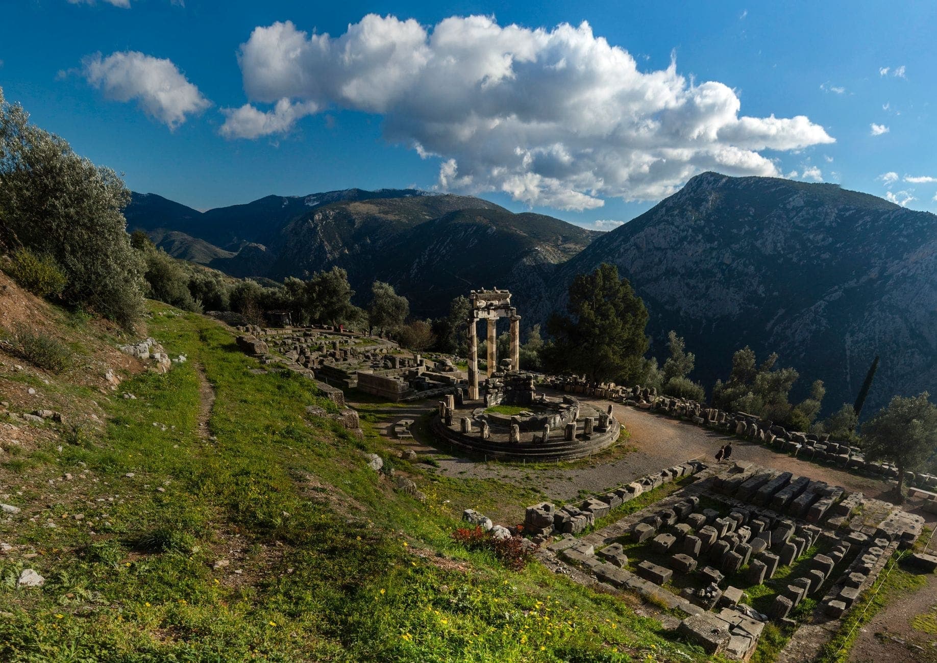 Panoramic view of the Delphi archaeological site with the Tholos of Delphi and ancient ruins surrounded by green mountains under a bright blue sky with clouds.