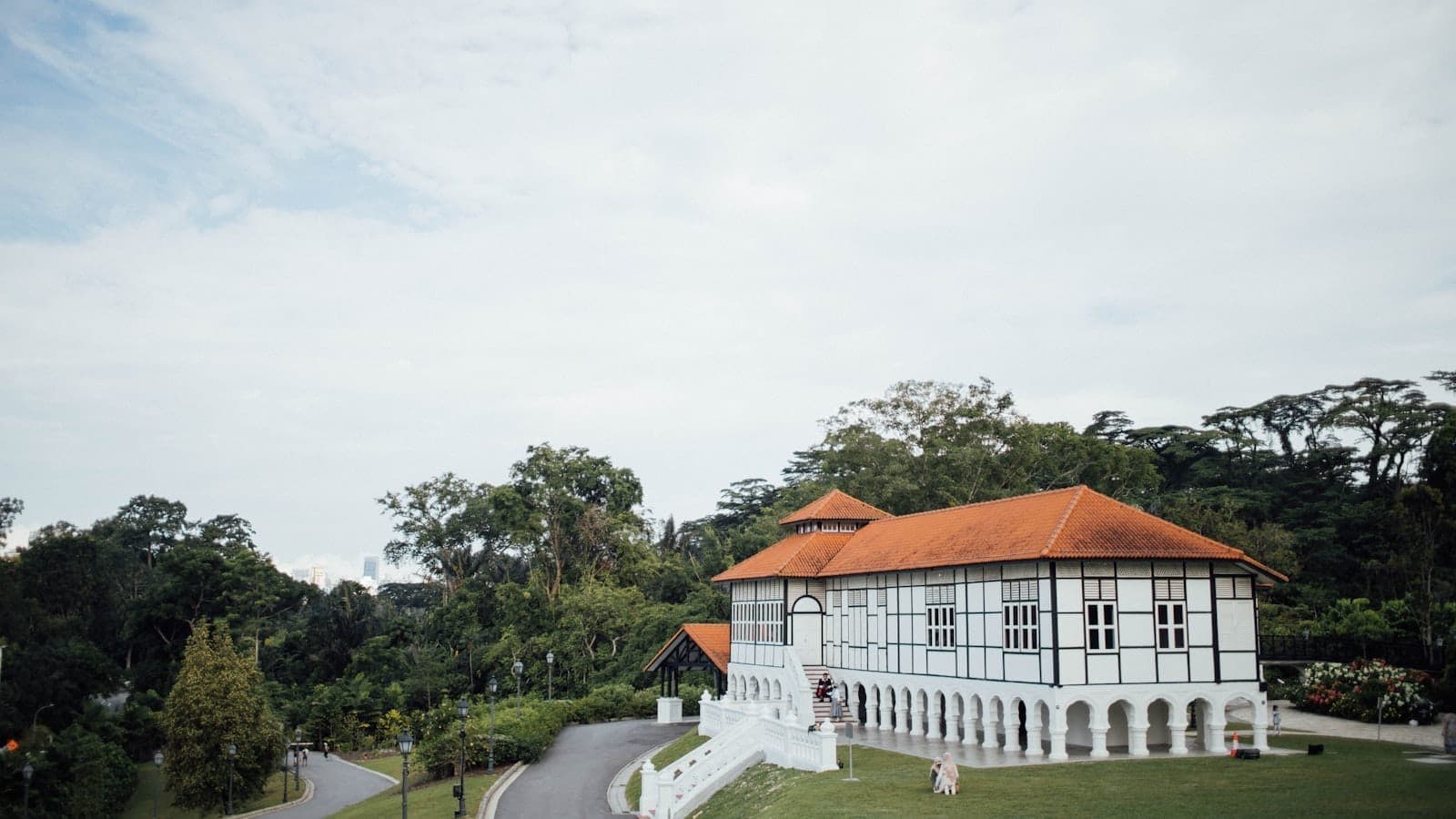 Wide view of a historic white colonial barracks with orange roof, surrounded by lush greenery and a winding driveway under a bright sky in Dempsey Hill, Singapore.