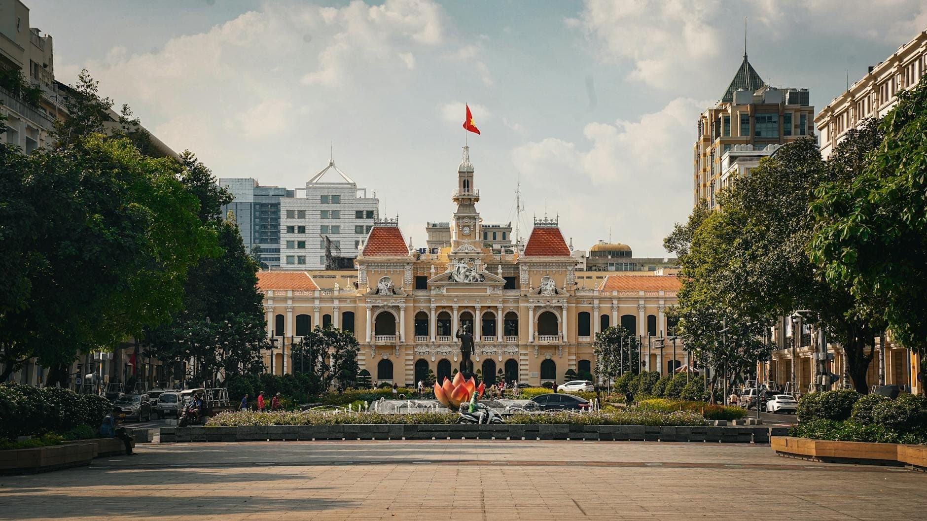 Malawak na tingin sa Ho Chi Minh City Hall na may maliwanag na asul na langit, napapaligiran ng mga puno, isang plasa na may palong-lotus, at mga modernong gusali sa likod.