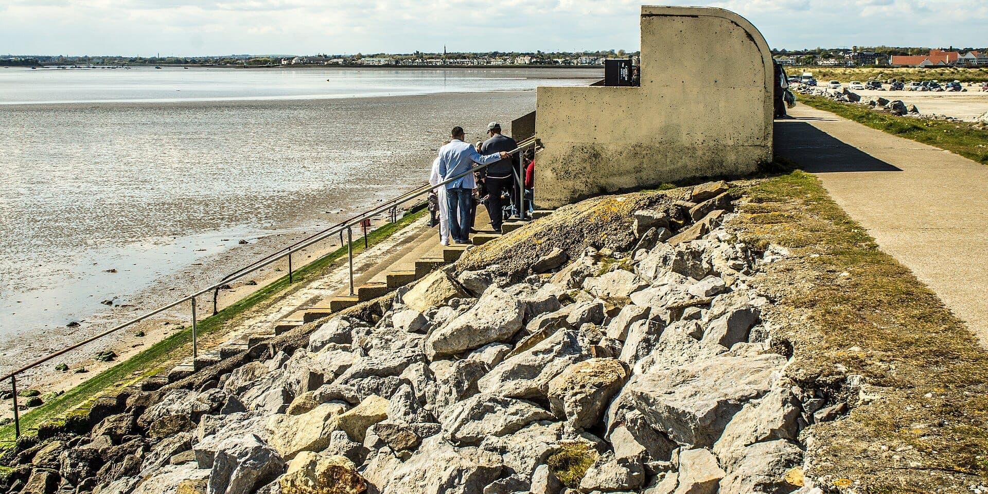 Besucher steigen betonierte Stufen vom Parkplatz hinunter auf den Dollymount Strand, mit den Wattflächen von Bull Island und den Häusern Dublins in der Ferne.