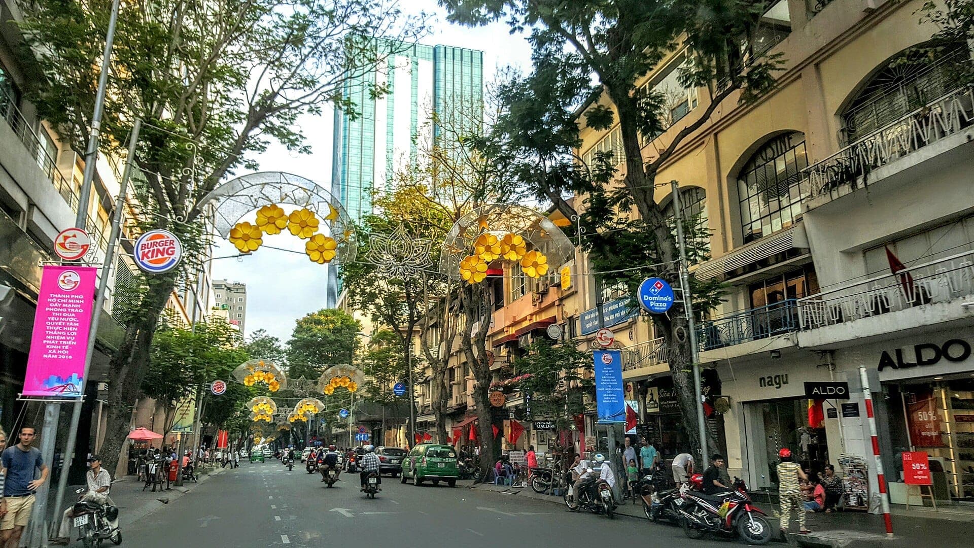 Cảnh đường phố trên đường Đồng Khởi tại Sài Gòn, hai bên là các tòa nhà kiến trúc thực dân, tòa nhà cao tầng, cửa hàng, đèn trang trí màu vàng và người đi bộ.