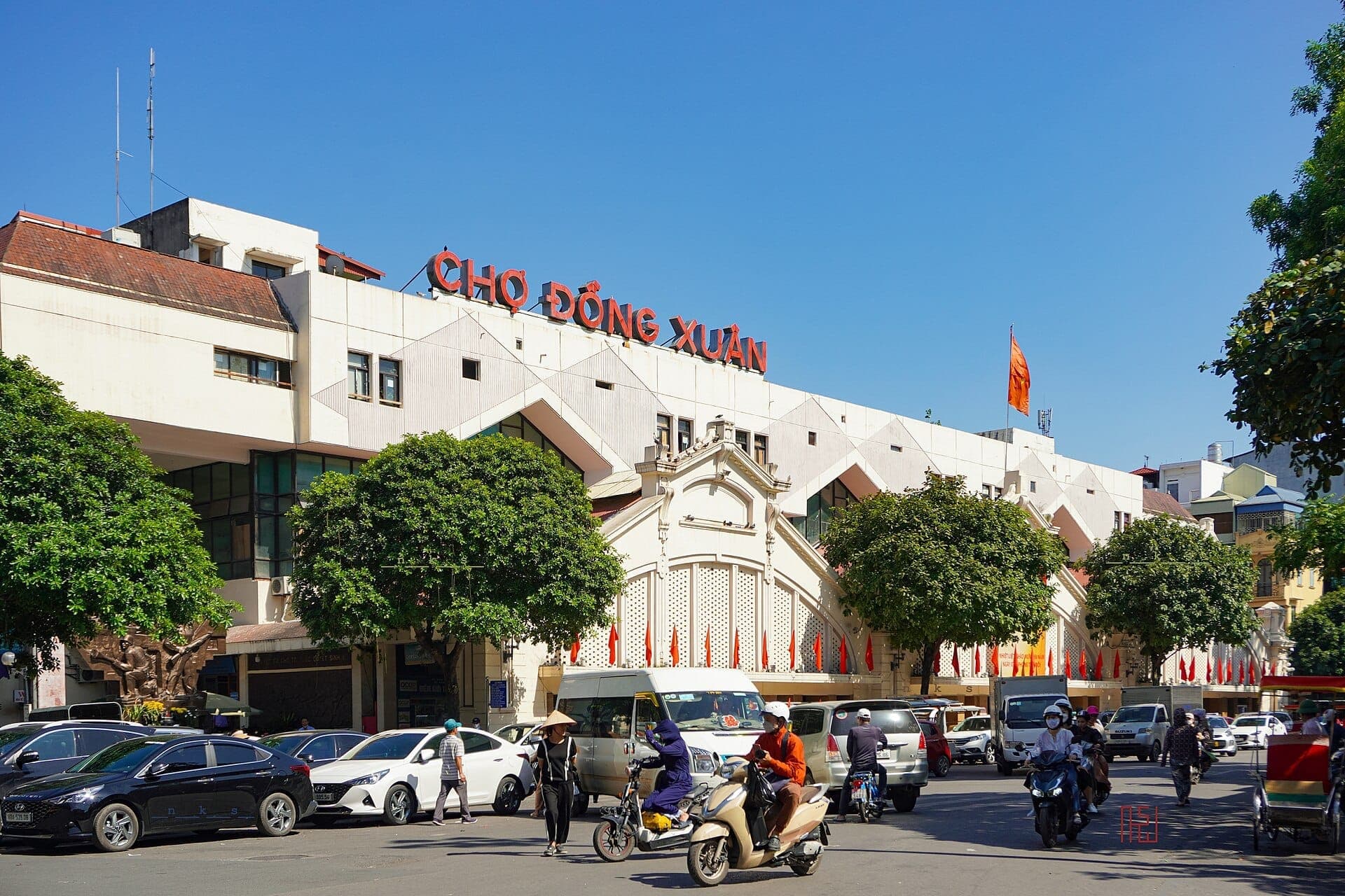 Cảnh bên ngoài chợ Đồng Xuân tại Hà Nội, nổi bật với mặt tiền màu trắng, biển hiệu đỏ và dòng xe cộ tấp nập dưới bầu trời xanh trong.