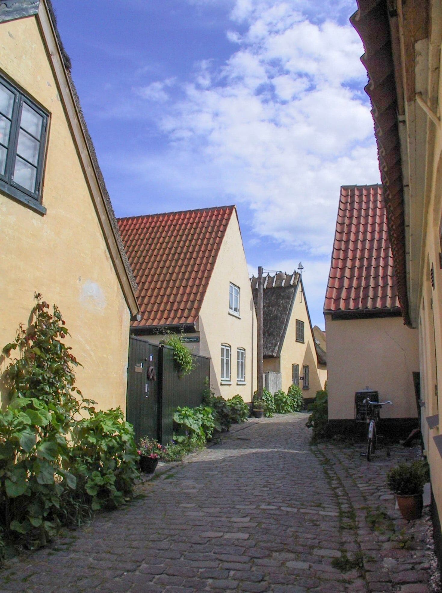 Case color ocra giallo con tetti di tegole rosse costeggiano una stretta strada acciottolata nella città vecchia di Dragør sotto un cielo blu parzialmente nuvoloso.