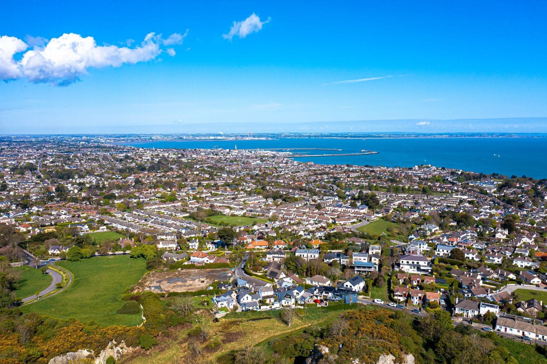 Weite Luftaufnahme eines Küstendorfes in der Nähe von Dublin mit Häusern, Grünflächen und dem Meer, das sich unter einem blauen Himmel in die Ferne erstreckt.