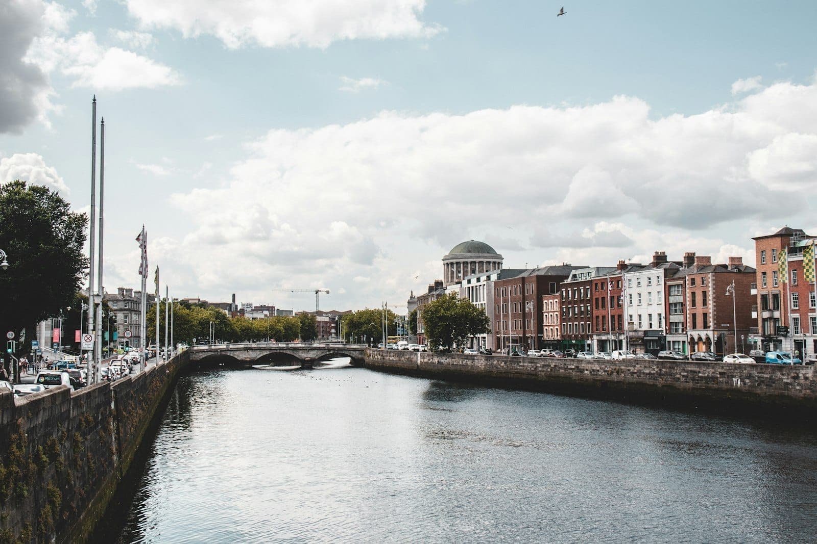 Heller Tagesblick auf Dublins Fluss Liffey mit klassischen georgianischen Gebäuden, einer Steinbrücke und der Kuppel der Four Courts unter einem teils bewölkten Himmel.