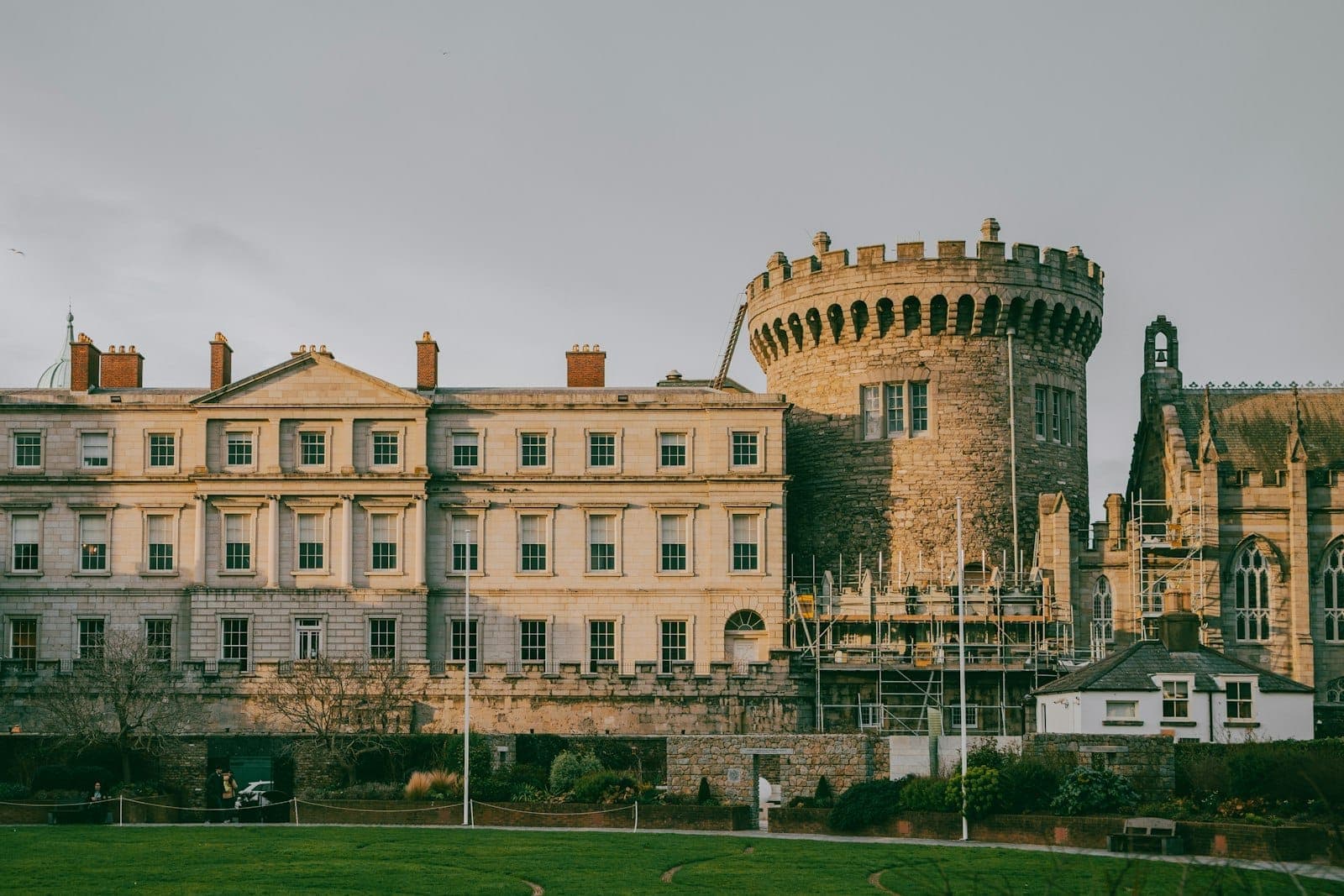 Wide landscape view of Dublin Castle featuring the medieval round tower, State Apartments, and adjoining green lawn under a cloudy sky.