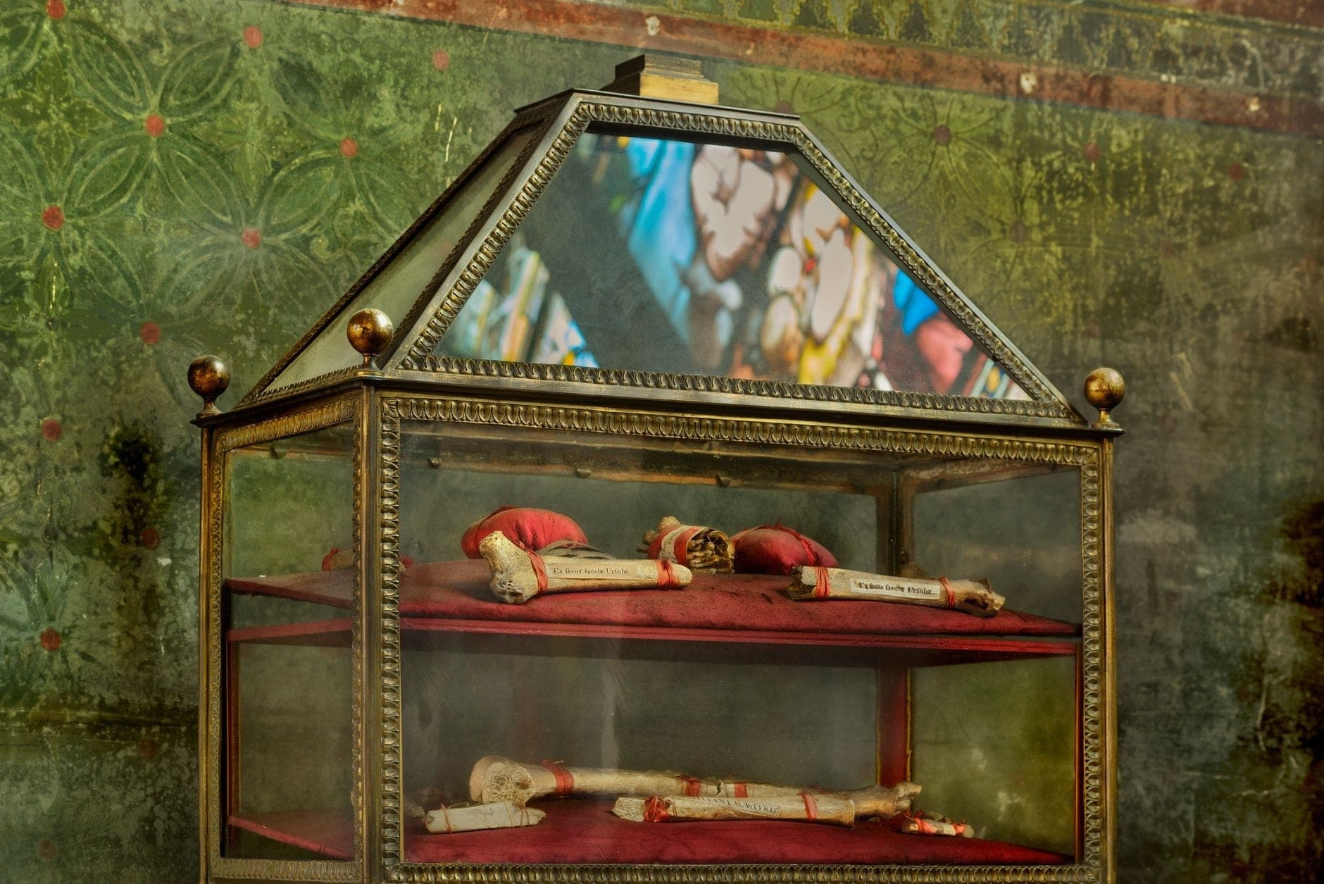 Glasreliquiar mit in rotes Tuch gehüllten Reliquien, mit einem Buntglasreflex dahinter, im Inneren einer Kirche.