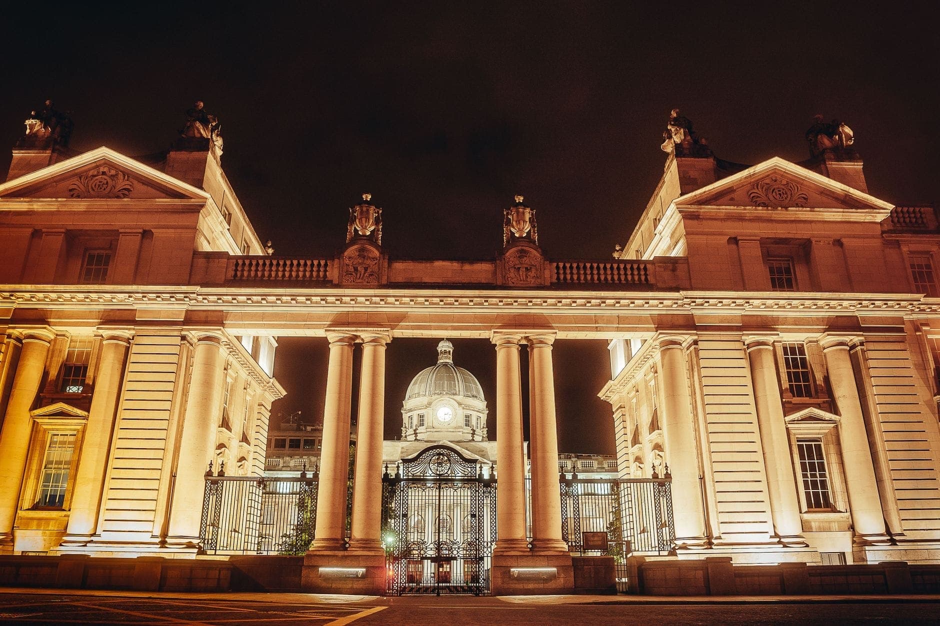 Cancelli neoclassici illuminati con colonne e edificio con cupola sullo sfondo, fotografati di notte nel centro di Dublino.