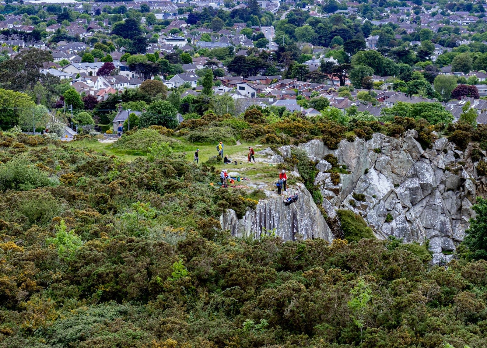 Vista su un affioramento roccioso con persone che camminano e si arrampicano, circondato da verde, con i quartieri residenziali di Dublino sullo sfondo.