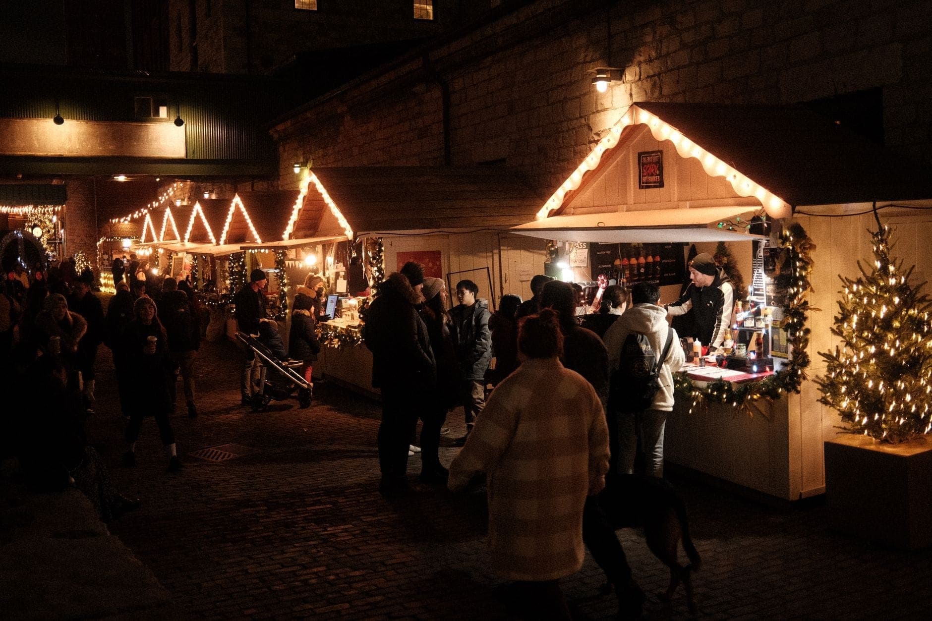 Menschenmassen stöbern an beleuchteten Holzständen auf einem Weihnachtsmarkt im Freien am Abend, mit festlichen Dekorationen und einer gemütlichen Winteratmosphäre.