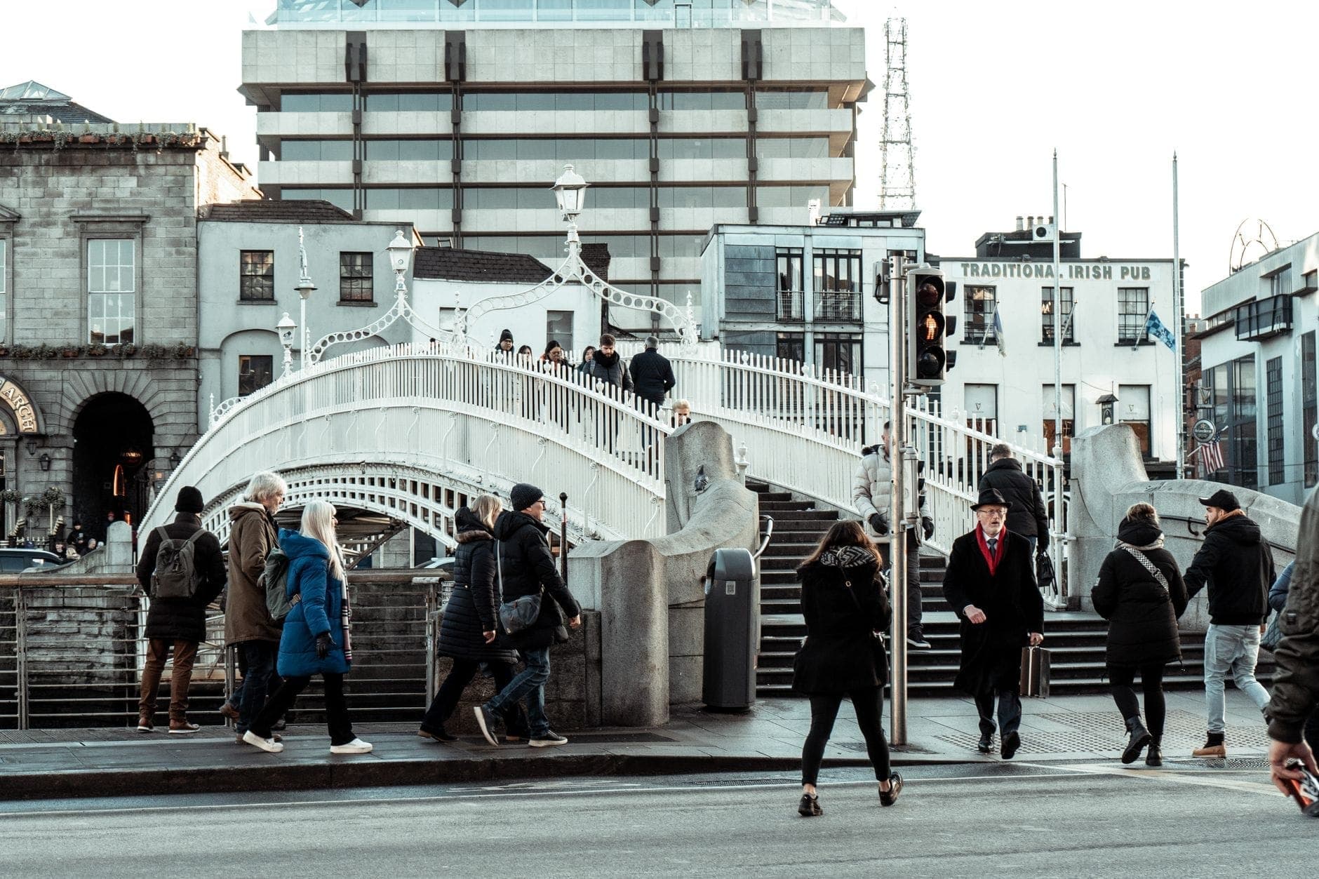 Menschen in Wintermänteln überqueren die Straße in der Nähe der ikonischen Ha'penny Bridge in Dublin an einem bedeckten Tag.