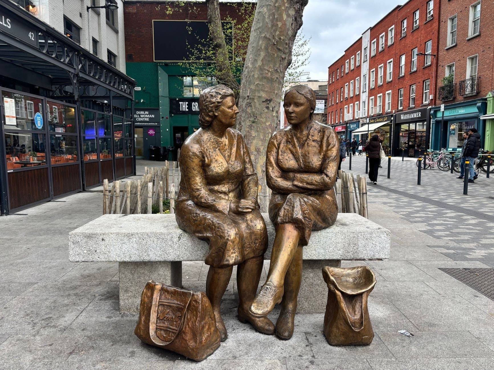Estátua de bronze de duas mulheres sentadas num banco numa movimentada rua de Dublin com lojas e edifícios de tijolo vermelho.