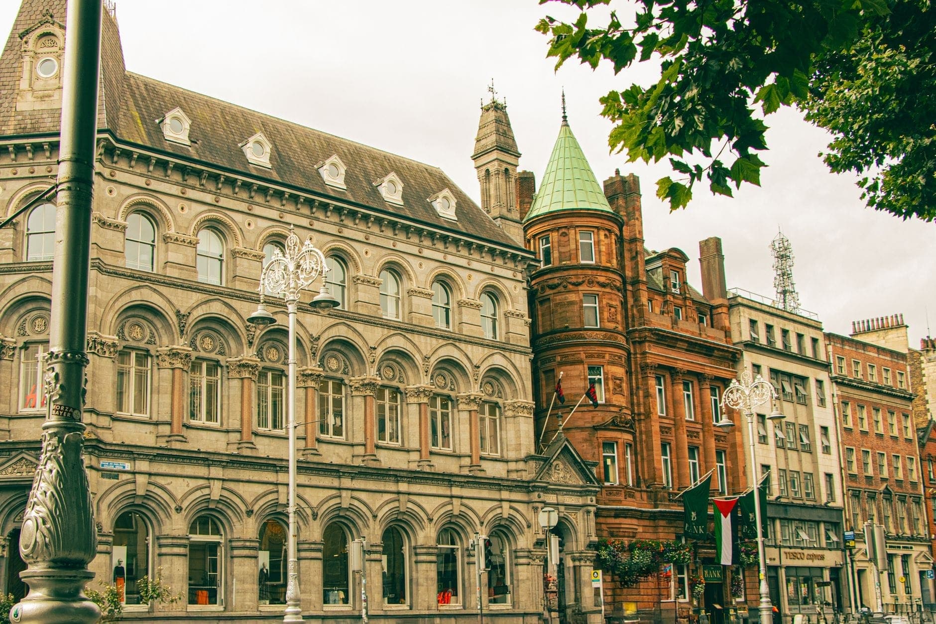 Elegantes edifícios históricos ao longo de uma rua de Dublin com fachadas ornamentadas e candeeiros de rua, árvore frondosa acima, mostrando a arquitetura do centro da cidade.