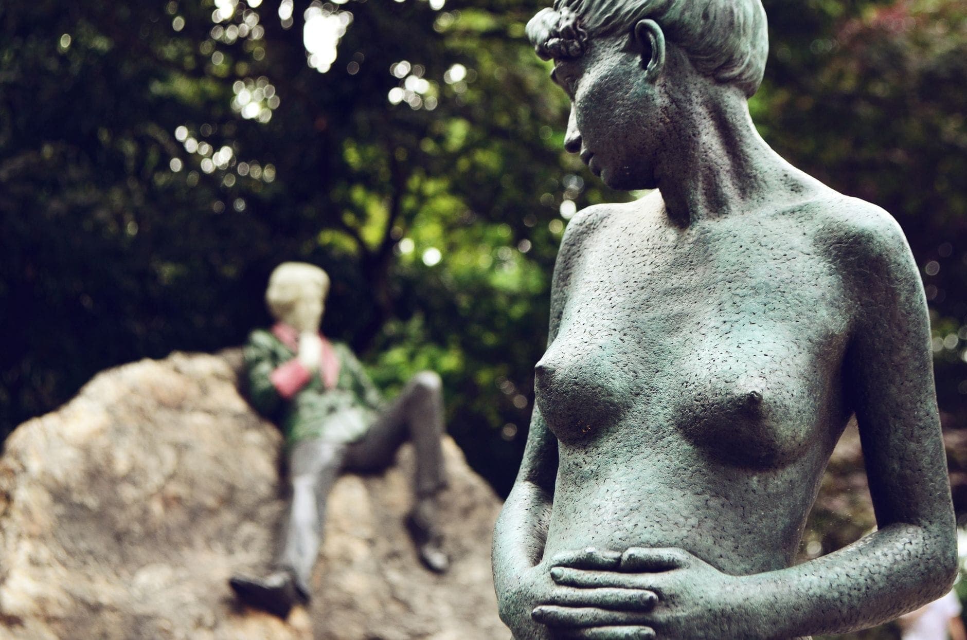 Estátua de bronze de uma mulher em primeiro plano, com a colorida estátua reclinada de Oscar Wilde visível sobre uma rocha em um parque de Dublin.
