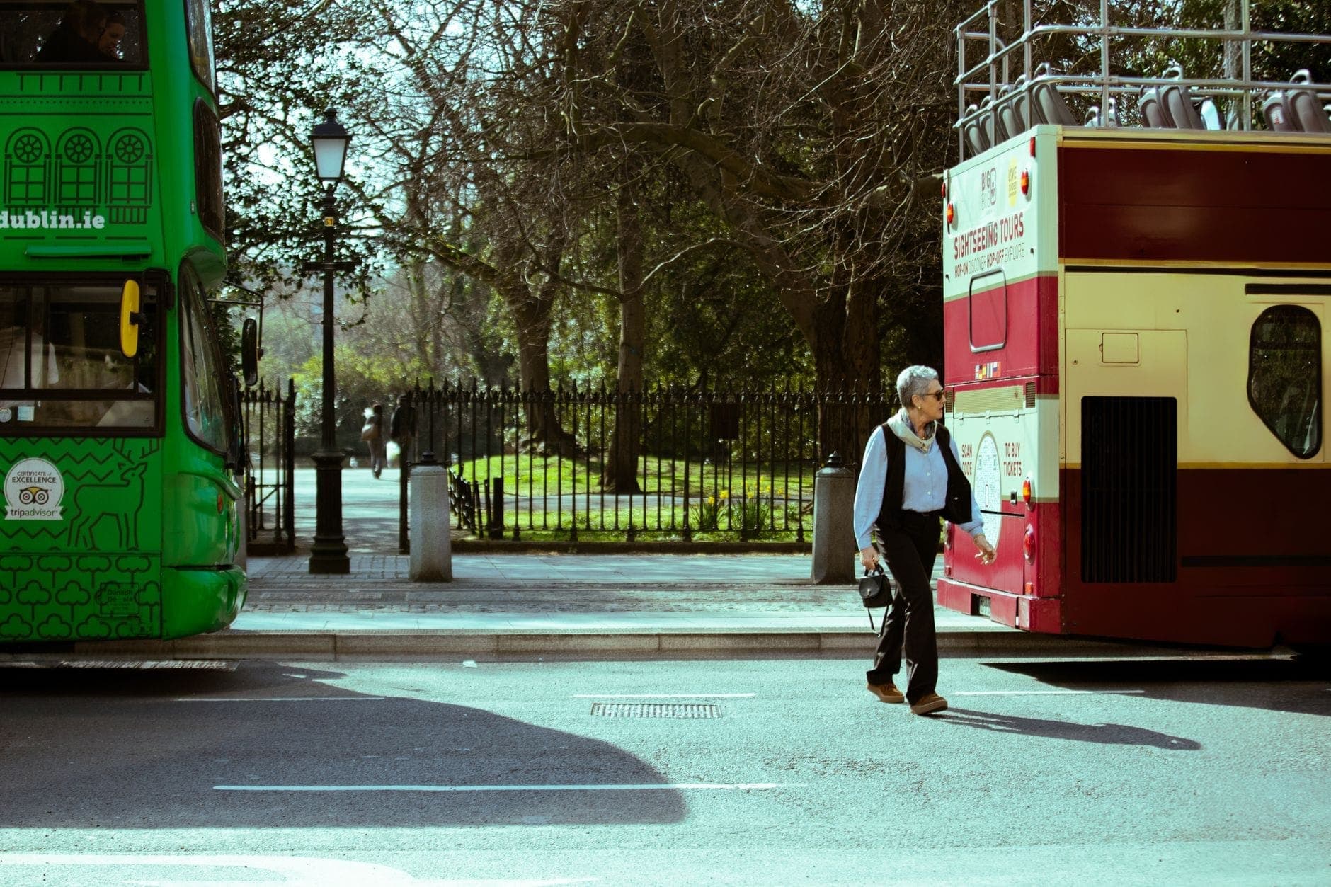 Homem a caminhar entre dois ônibus turísticos numa rua do centro de Dublin, com árvores e grade de parque ao fundo.