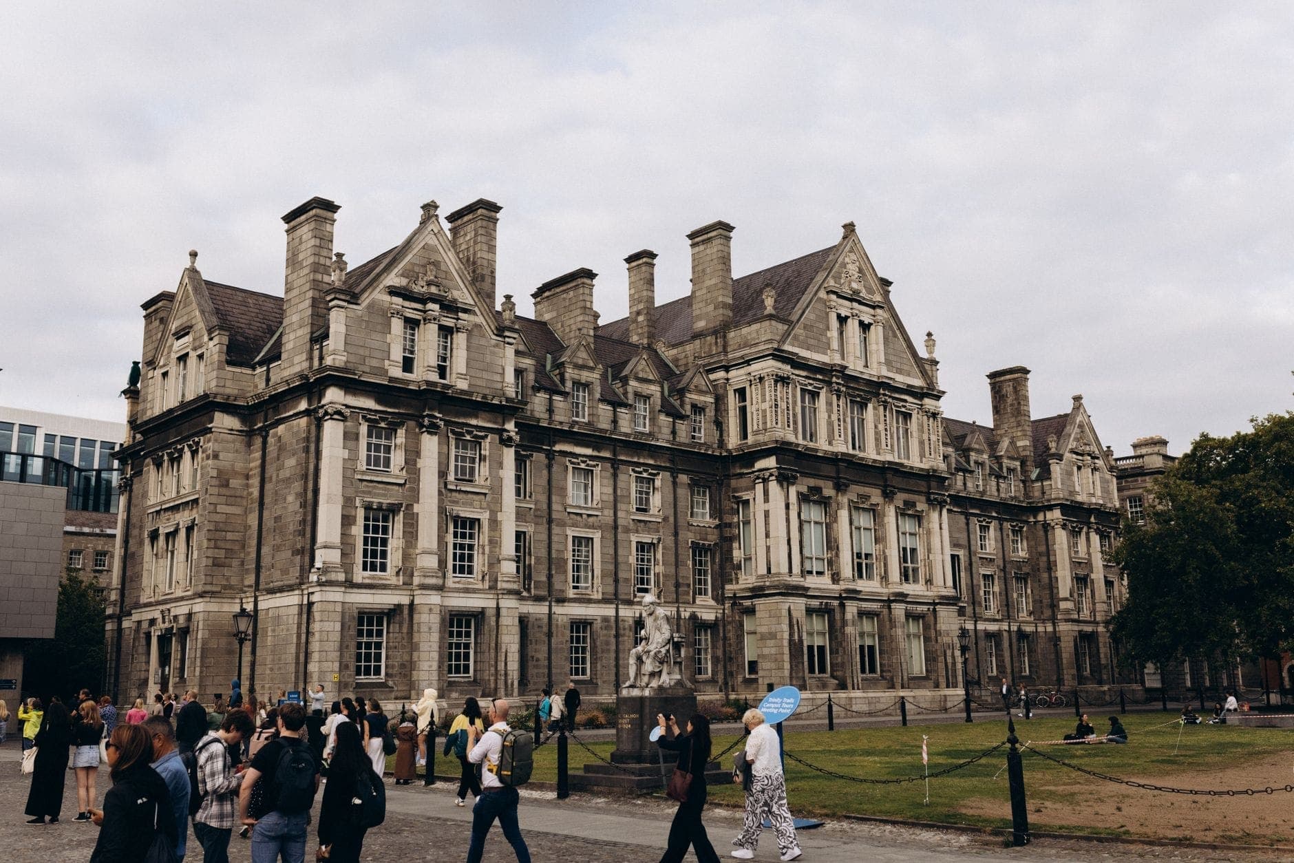 Gruppi di persone che camminano davanti a un imponente edificio storico al Trinity College Dublin in una giornata nuvolosa.