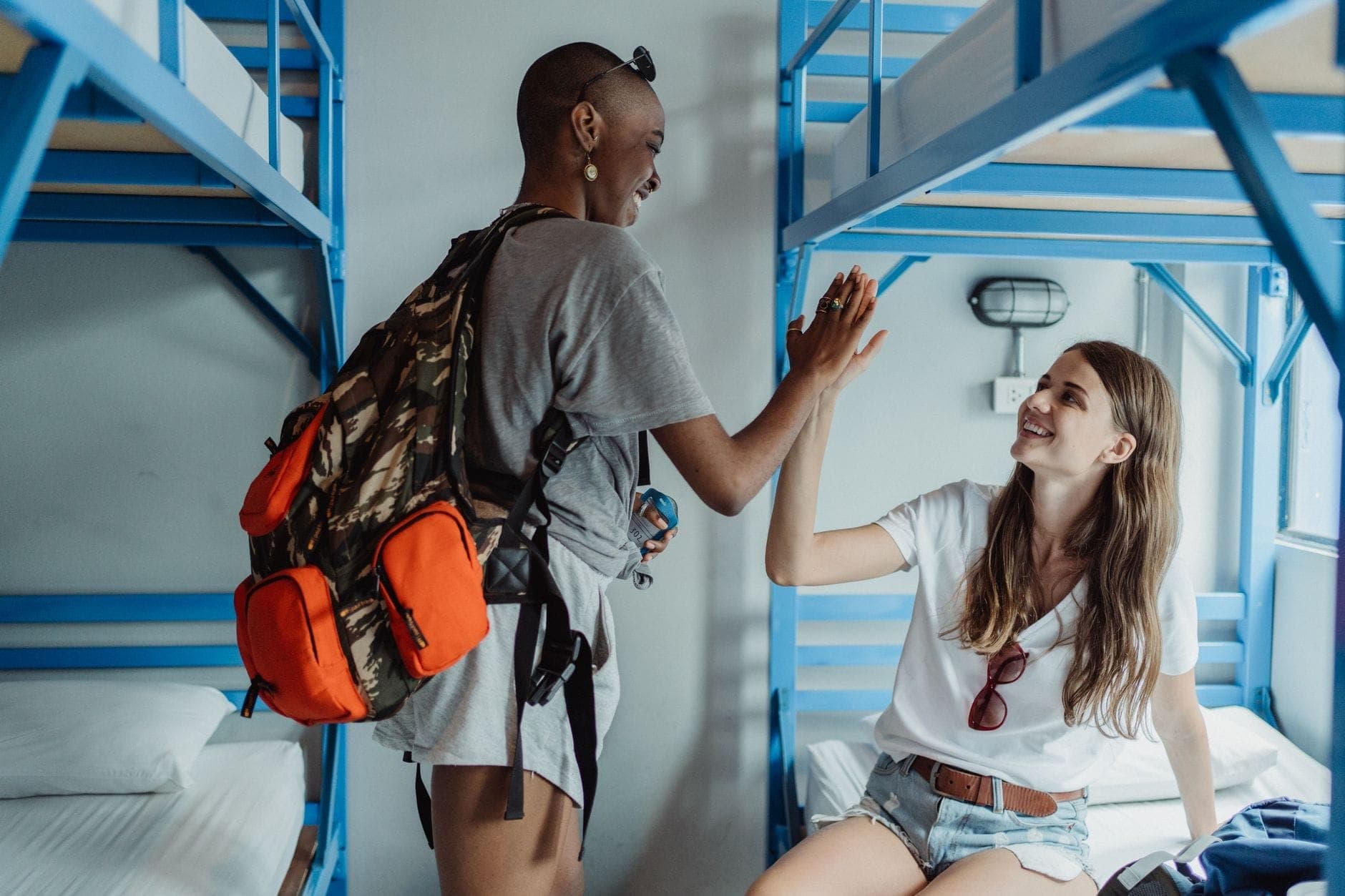 Due viaggiatori in una vivace stanza di ostello con letti a castello in metallo blu, uno seduto con uno zaino e uno che fa un high five.