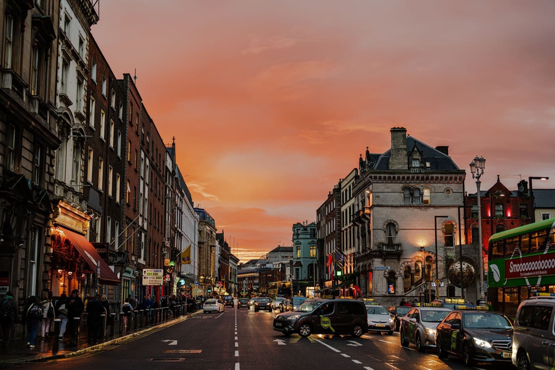 Vista ampia di una vivace strada di Dublino al tramonto, con edifici storici, pub, gente a passeggio e traffico sotto un cielo colorato.