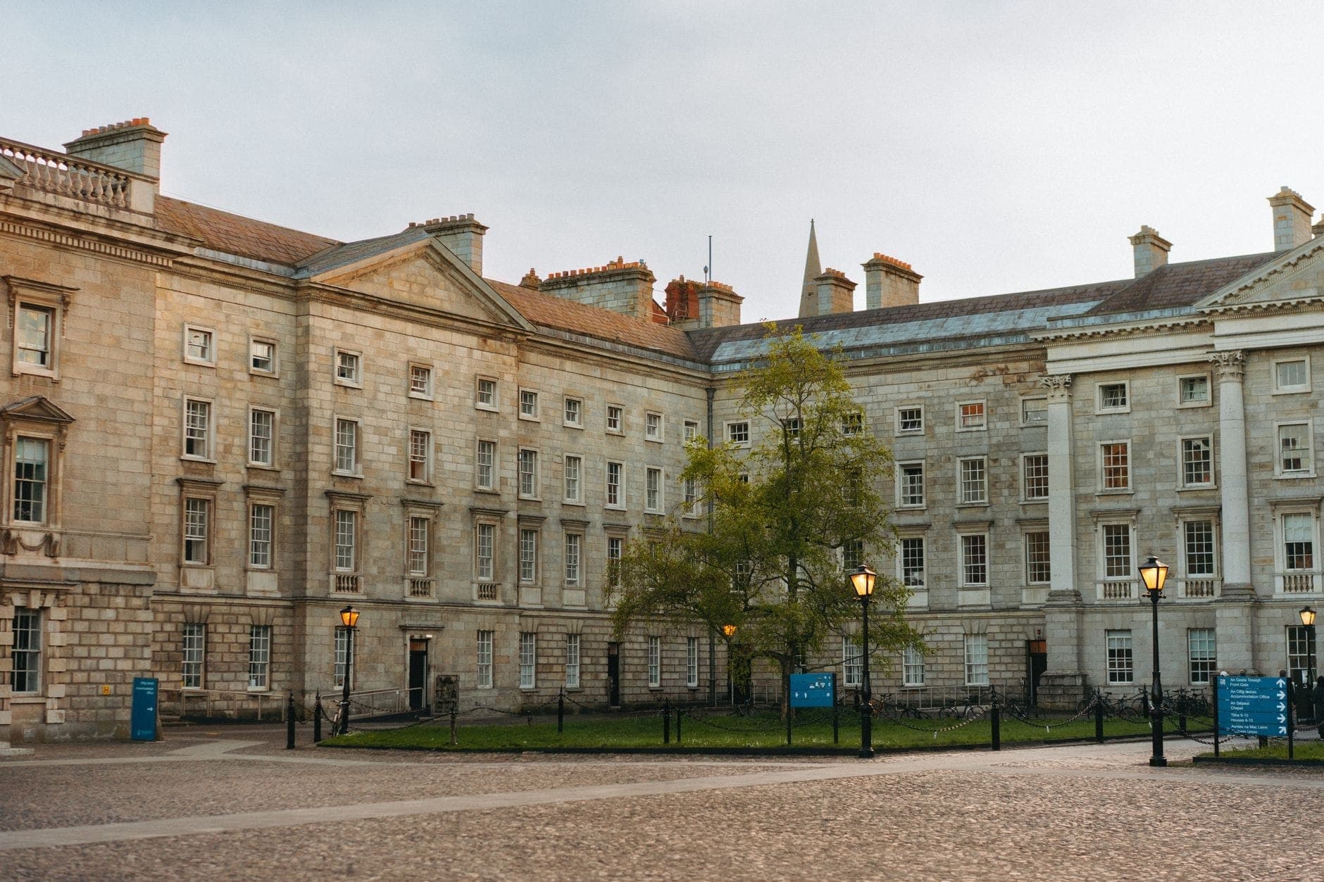 Ripresa grandangolare del cortile del Trinity College Dublin con storici edifici in pietra, un albero verde e cartelli informativi blu visibili in una giornata luminosa.