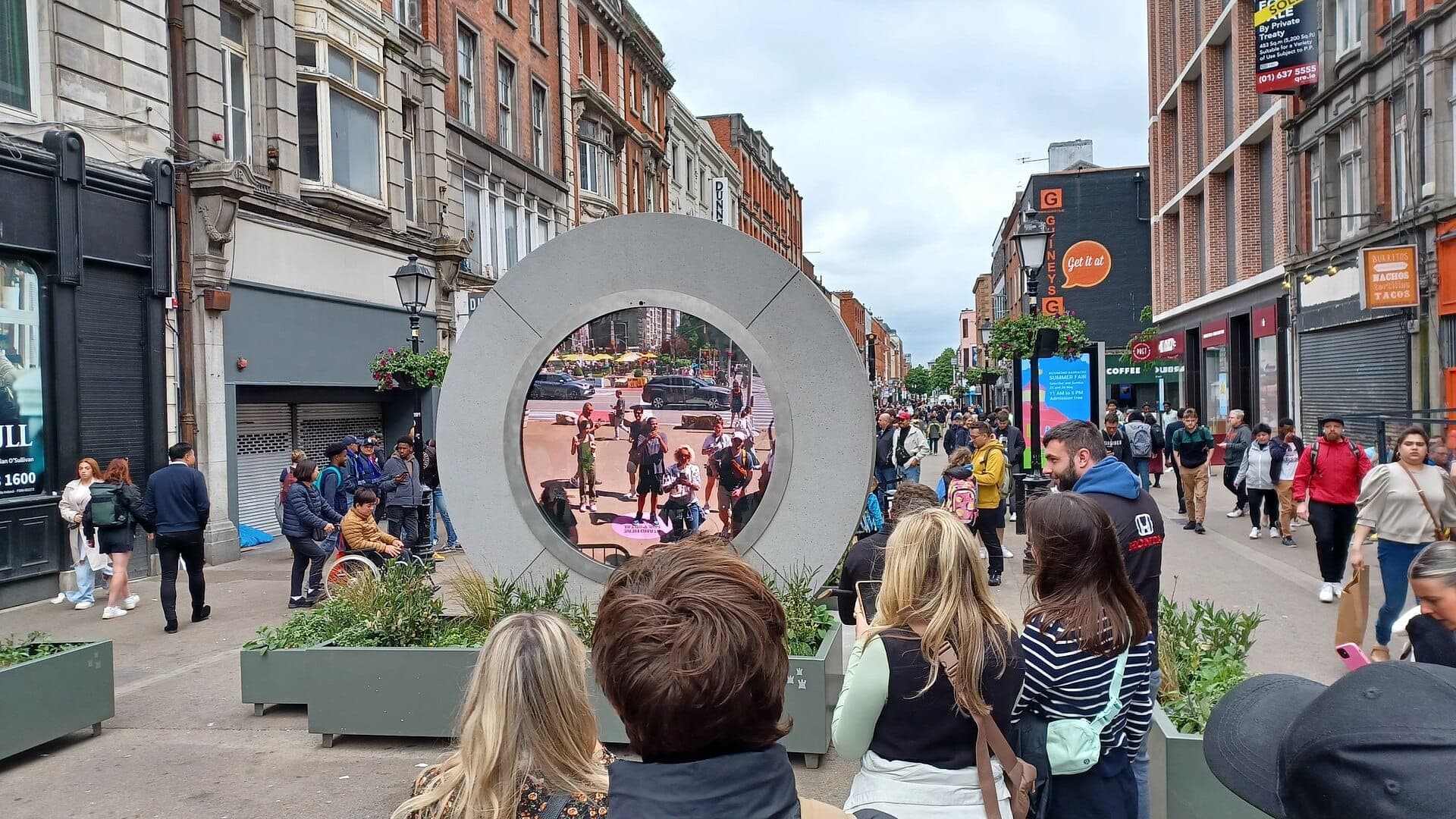 Nagtitipon ang mga tao sa O'Connell Street upang panoorin ang bilog na eskultura ng Dublin Portal na naglivestream ng buhay-lungsod mula sa ibang bansa sa isang makulimlim at maingay na araw.