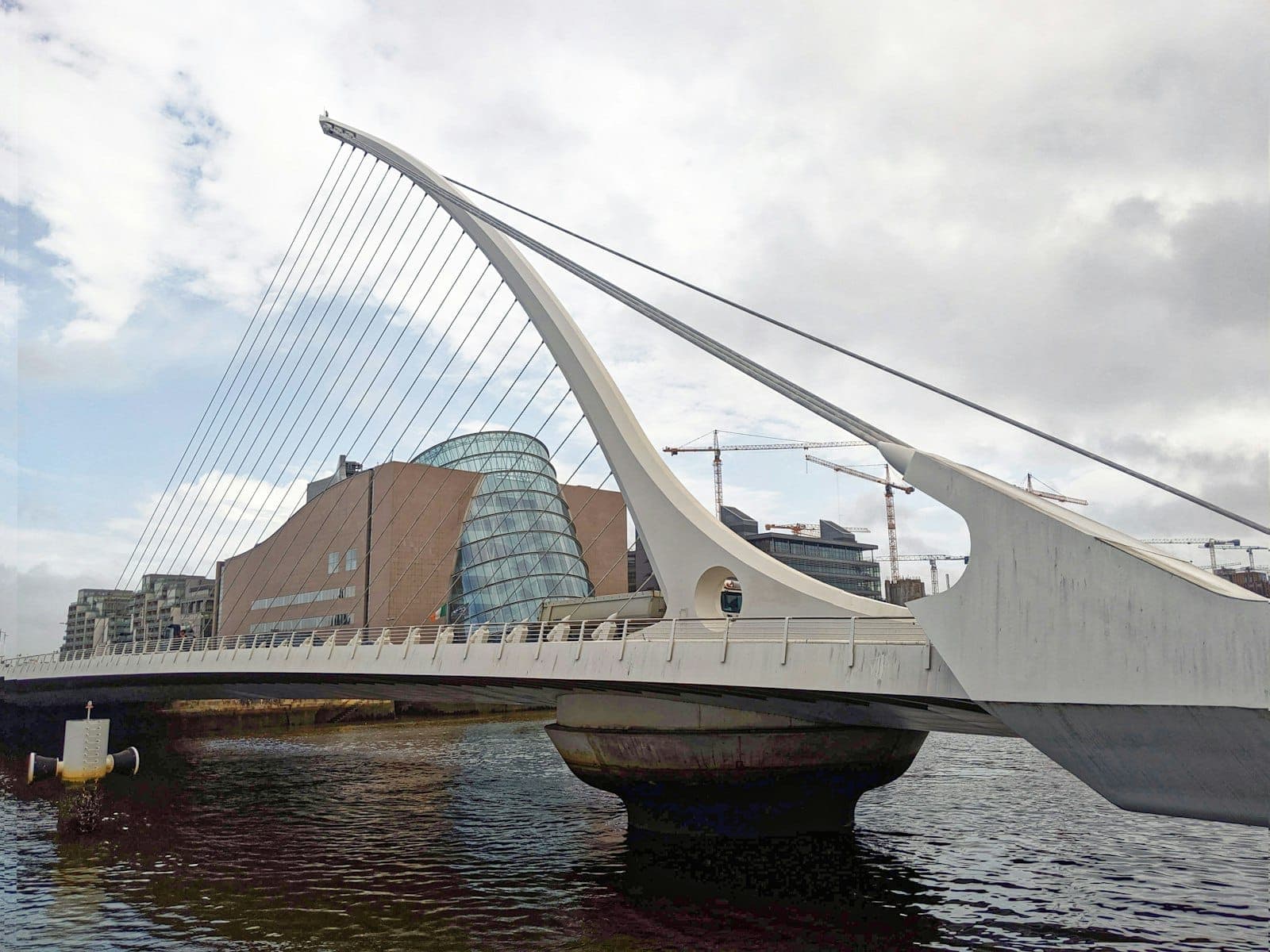 Samuel Beckett Bridge spanning the River Liffey in Dublin, with the Convention Centre and modern cityscape in the background under cloudy skies.