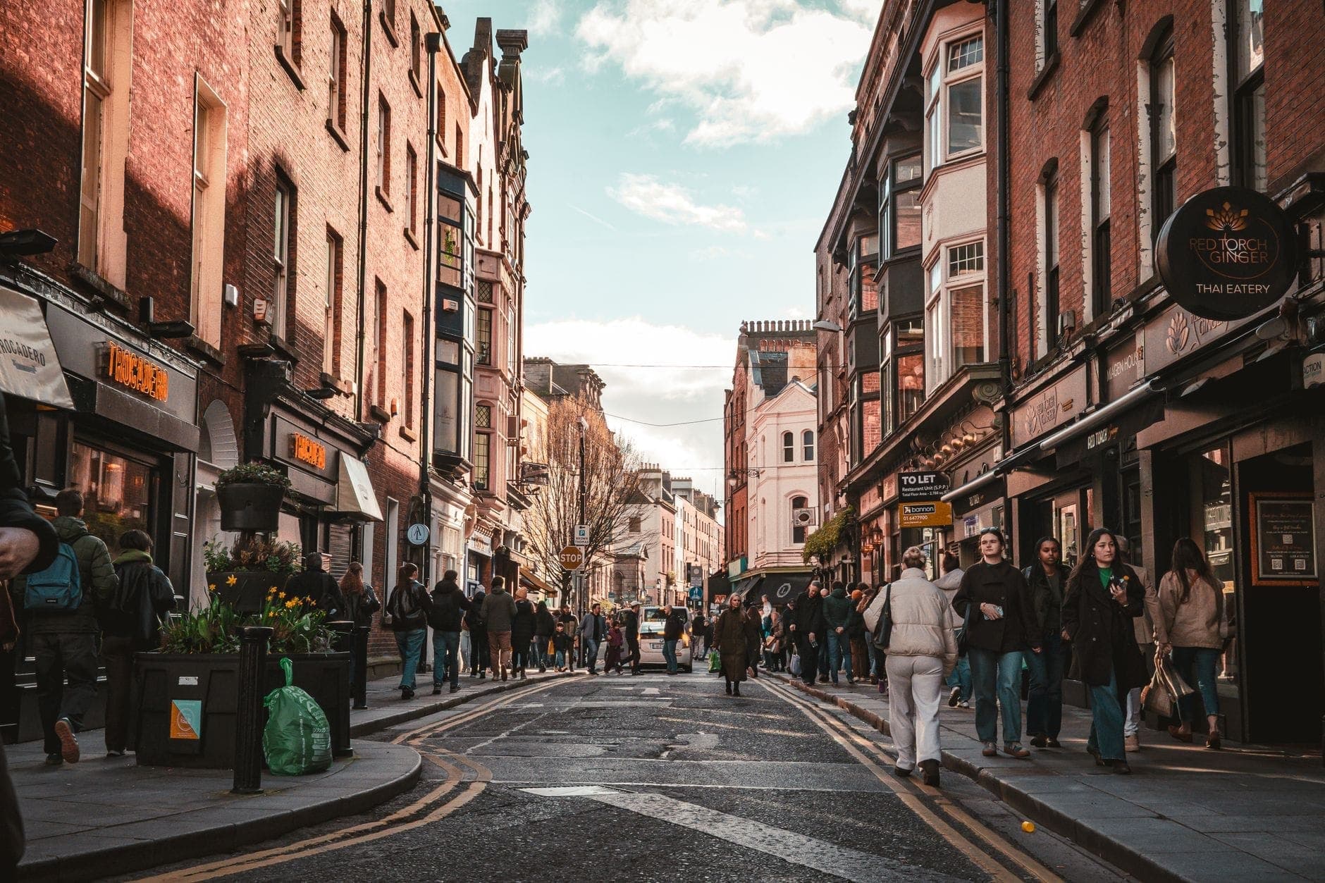 Belebte Einkaufsstraße in Dublin mit roten Backsteingebäuden, Läden und Cafés, gefüllt mit Passanten unter leicht bewölktem Himmel.