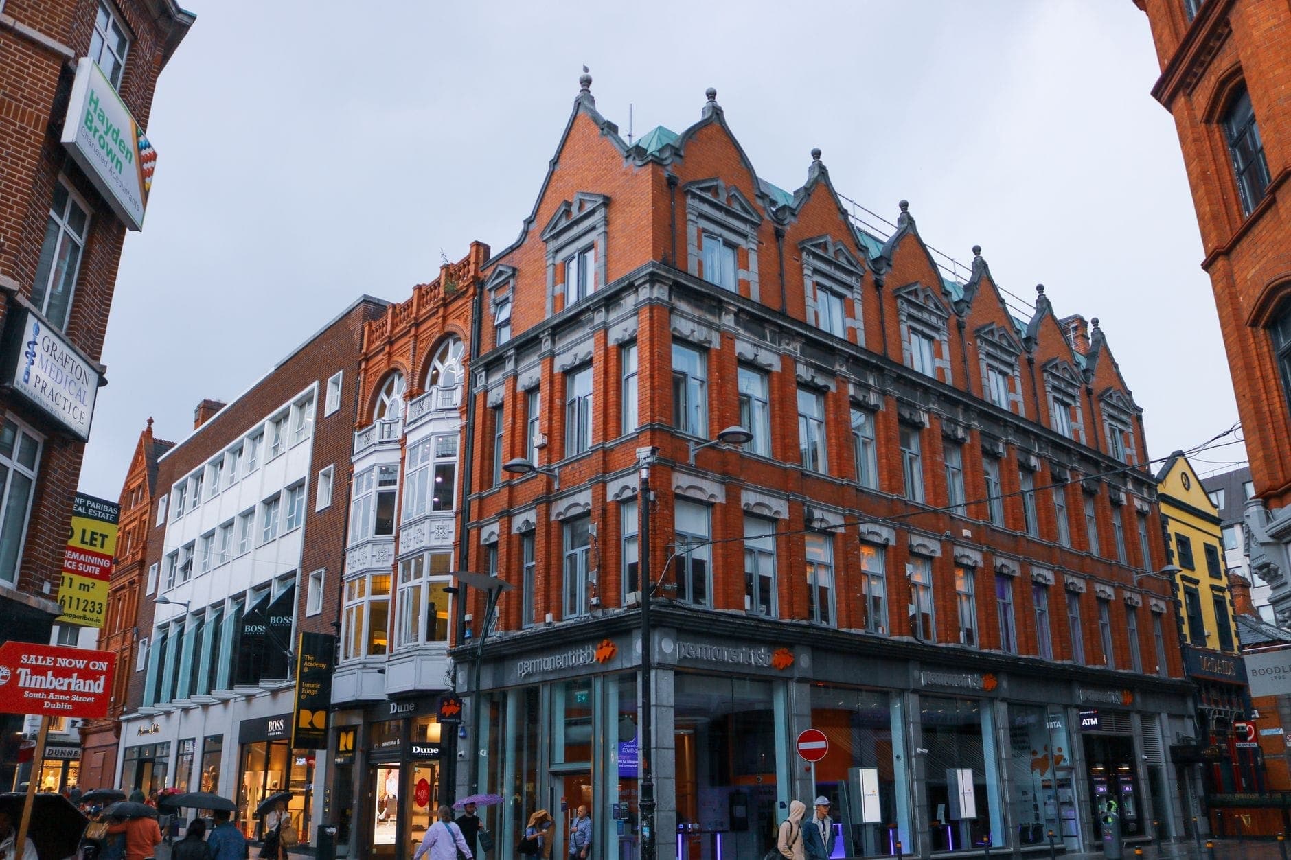 Großes, elegantes Backsteingebäude an einer belebten Fußgängerzone in Dublin mit Ladenschildern und vorbeigehenden Menschen.