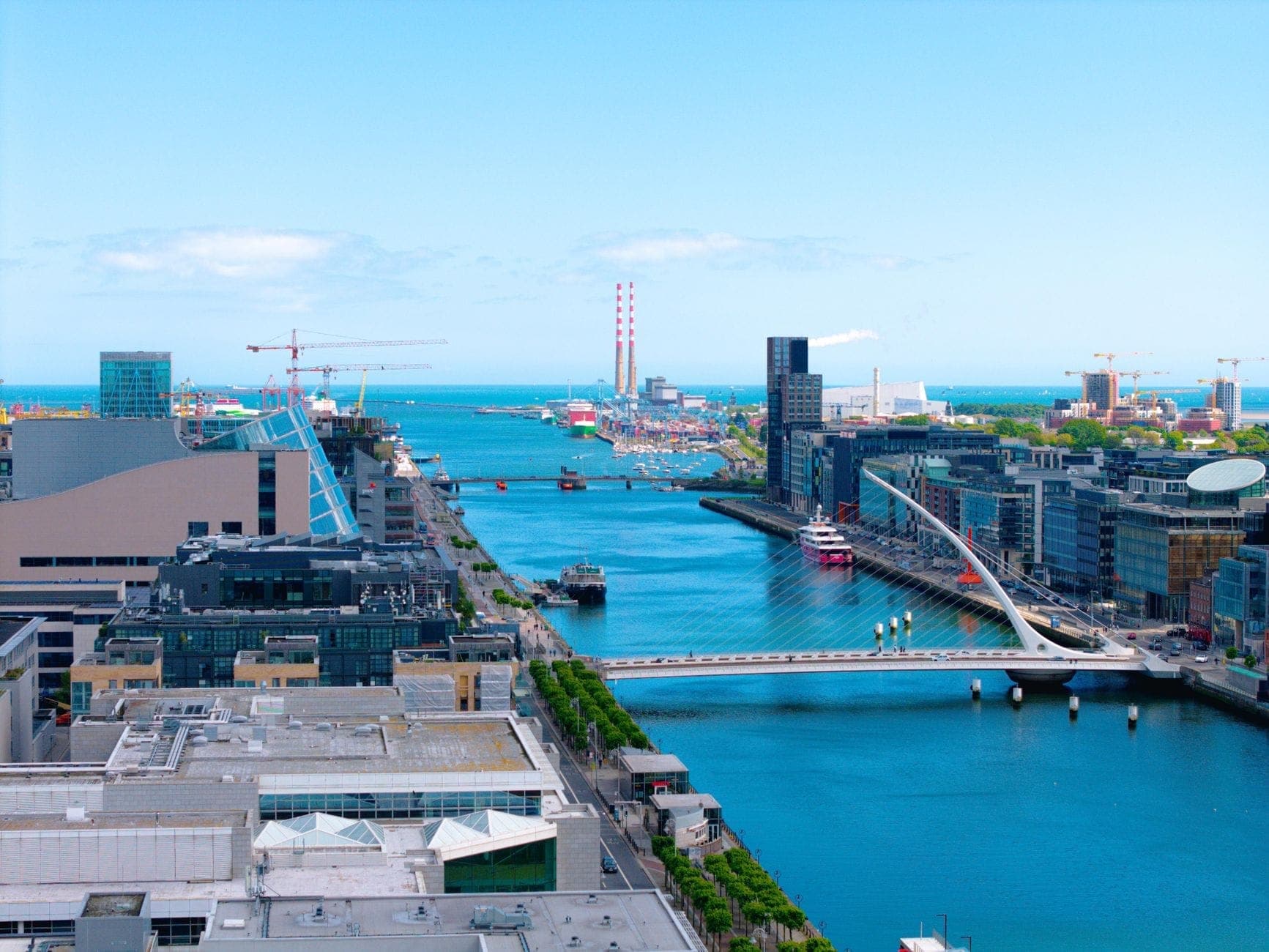 Vue aérienne des Dublin Docklands avec le Samuel Beckett Bridge, des bâtiments modernes et la River Liffey sous un ciel bleu vif.