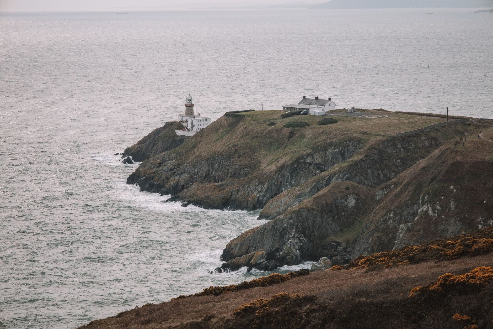 Promontoire côtier avec un phare et un cottage surplombant la mer, falaises spectaculaires et vagues, typique des promenades côtières de Howth à Dublin.