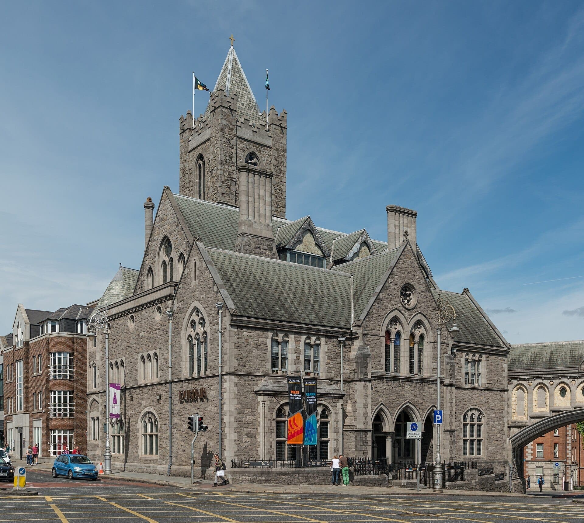 Widok zewnętrzny muzeum Dublinia, mieszczącego się w okazałym budynku w stylu neogotyckim, z kamiennymi łukami, wieżą, kolorowymi chorągwiami i bezchmurnym błękitnym niebem.