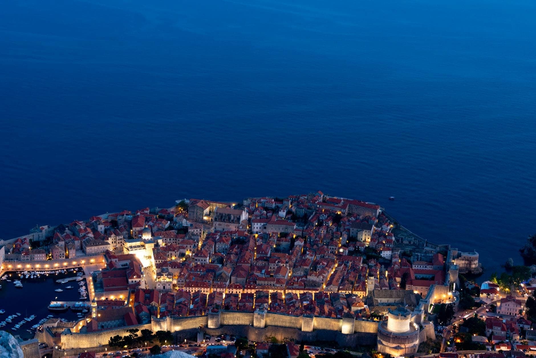 Cảnh hoàng hôn nhìn từ trên cao xuống khu phố cổ Dubrovnik với những bức tường pháo đài thời trung cổ được thắp sáng, phía dưới là mặt biển Adriatic phẳng lặng, gợi lên không khí sôi động của thành phố về đêm.