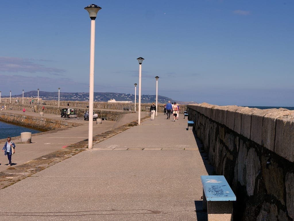 Weitblick auf den Ostpier von Dún Laoghaire mit Steinpromenade, Laternen, Bänken und Spaziergängern entlang der Küste unter klarem blauem Himmel.